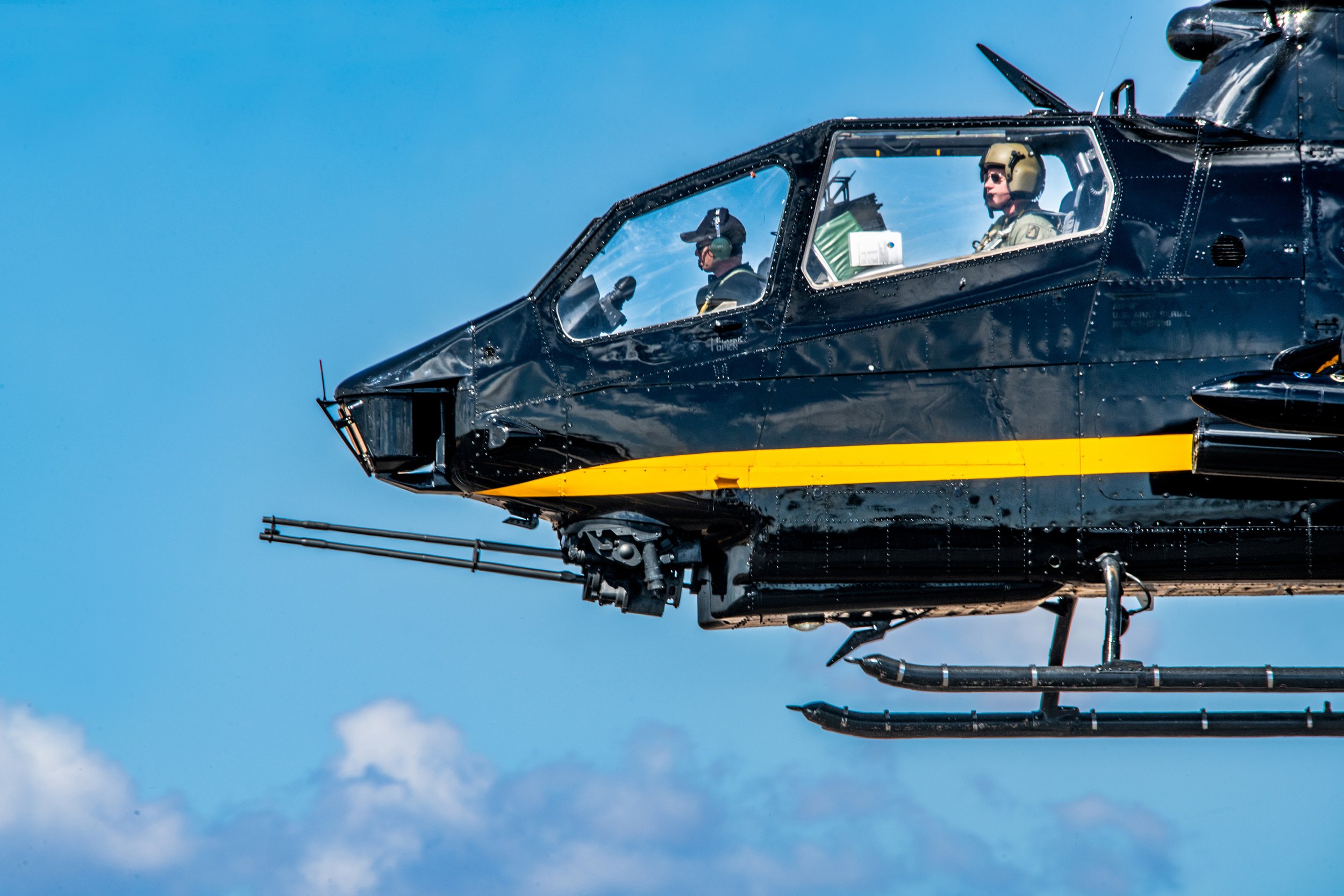 A black military helicopter in flight with two crew members in the cockpit, one wearing sunglasses and headset, the other with a hat, against a blue sky with clouds.