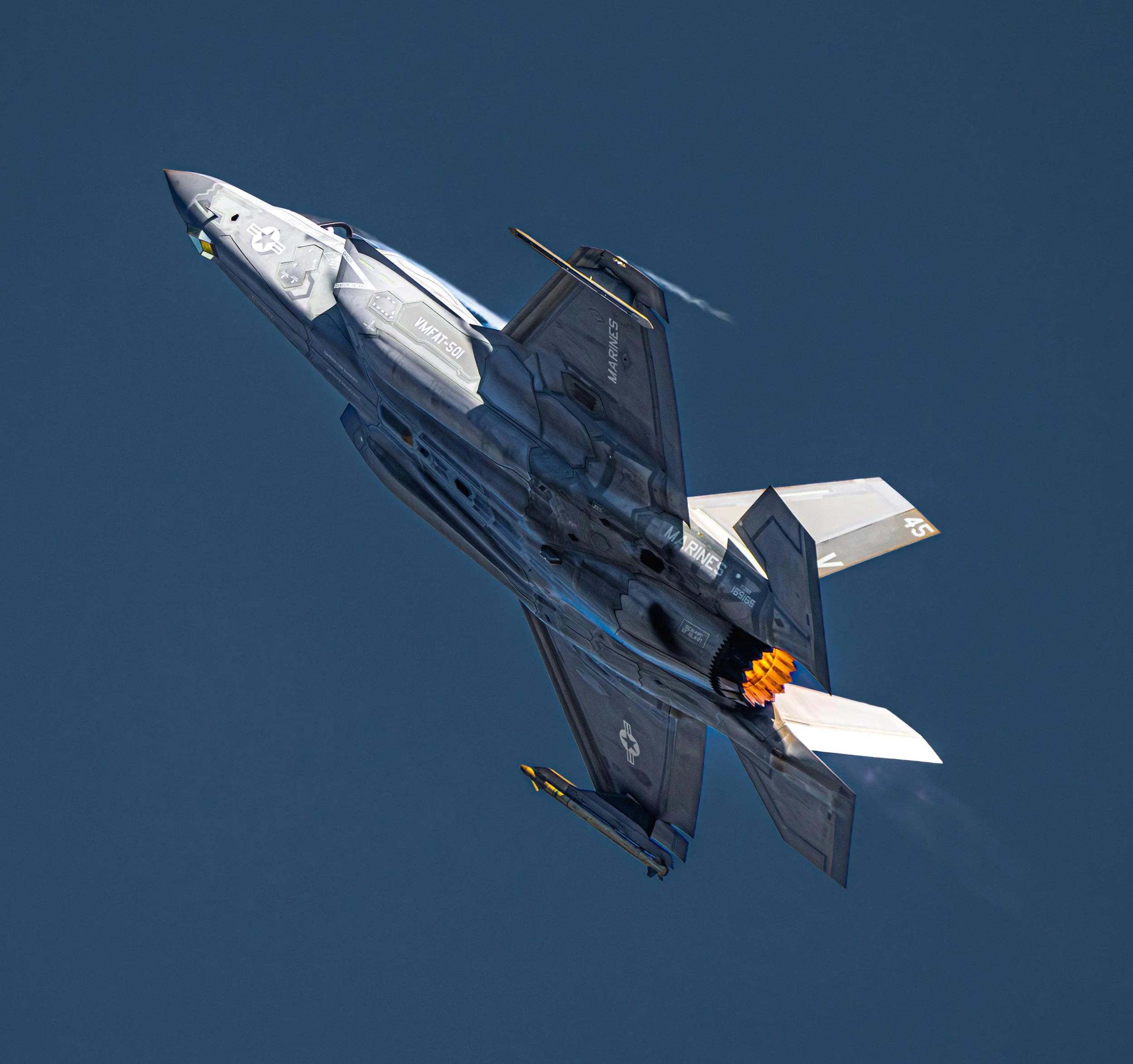 A fighter jet in flight against a clear blue sky, with afterburners ignited.