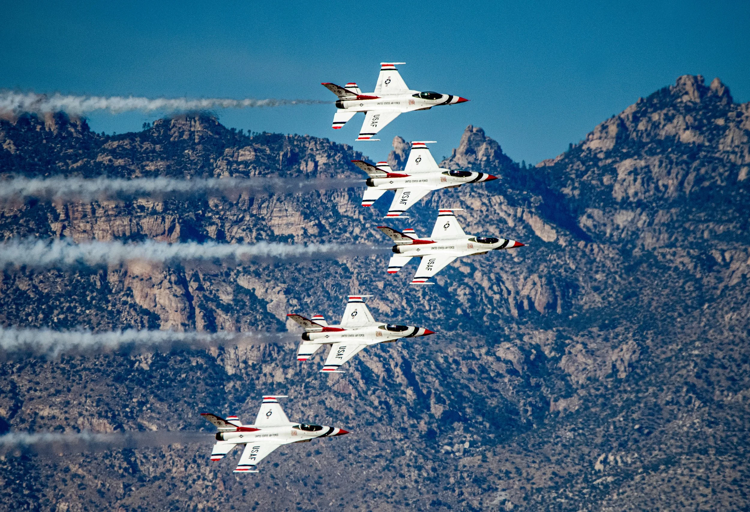 Five USAF fighter jets flying in close formation over rocky mountains, leaving white smoke trails in clear blue sky.