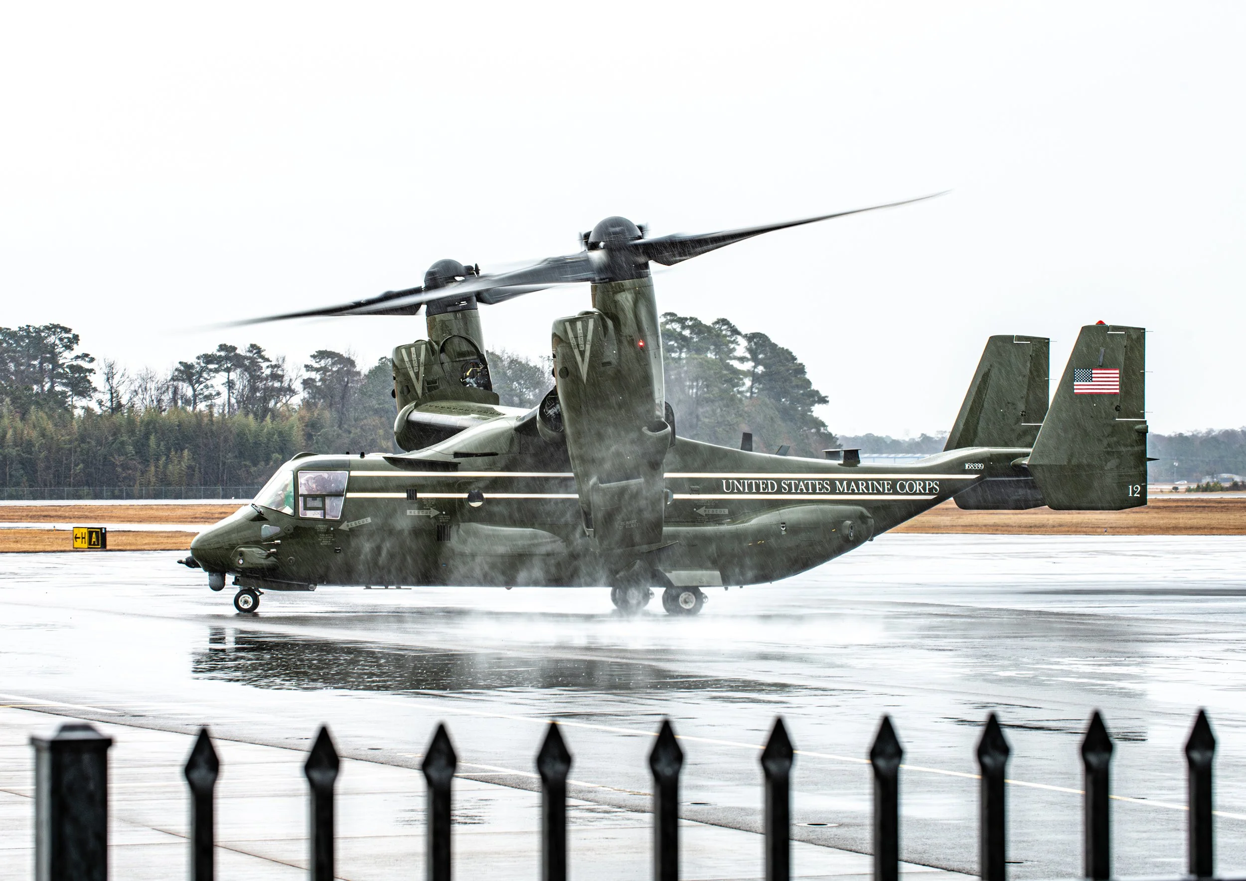 A military helicopter on a wet tarmac with water spraying from its blades, marked with 'United States Marine Corps' and an American flag.