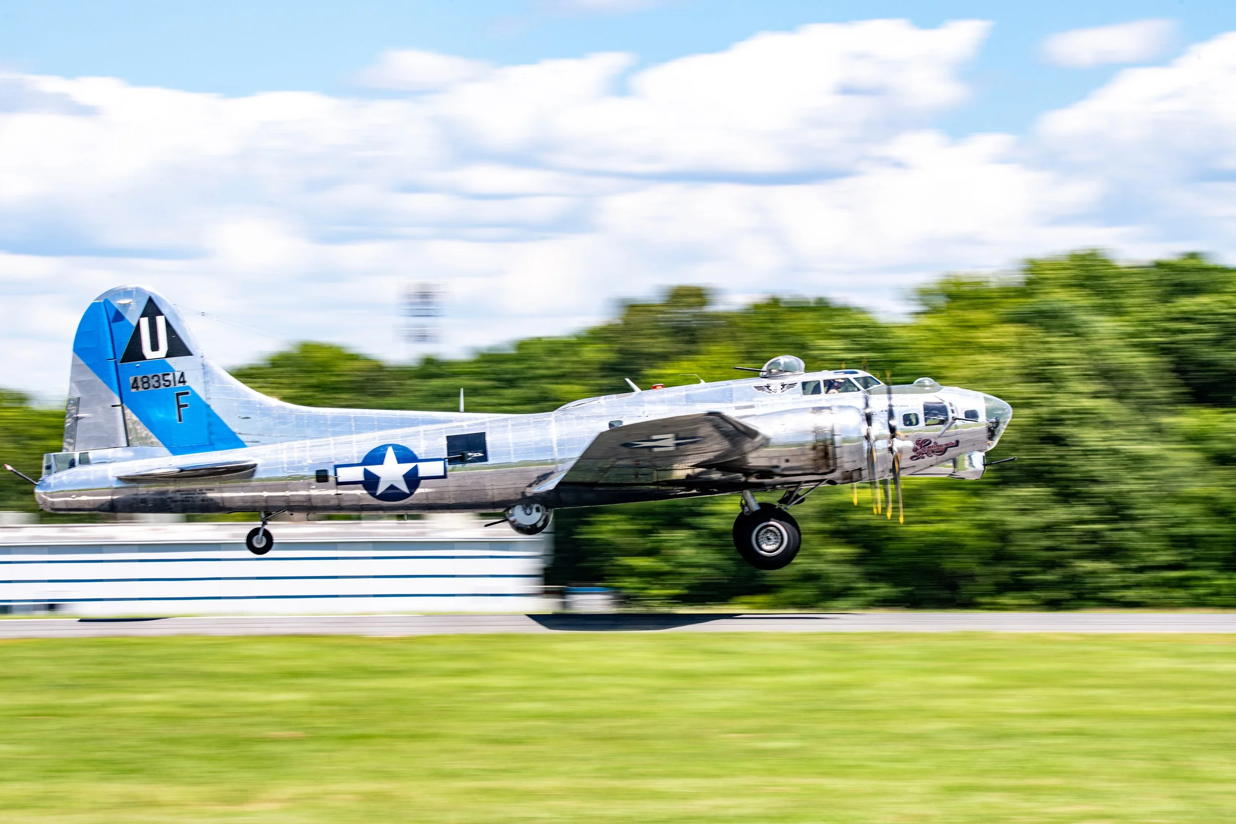 A vintage silver military airplane in mid-flight, flying low over a grassy airstrip with trees and partly cloudy sky in the background.