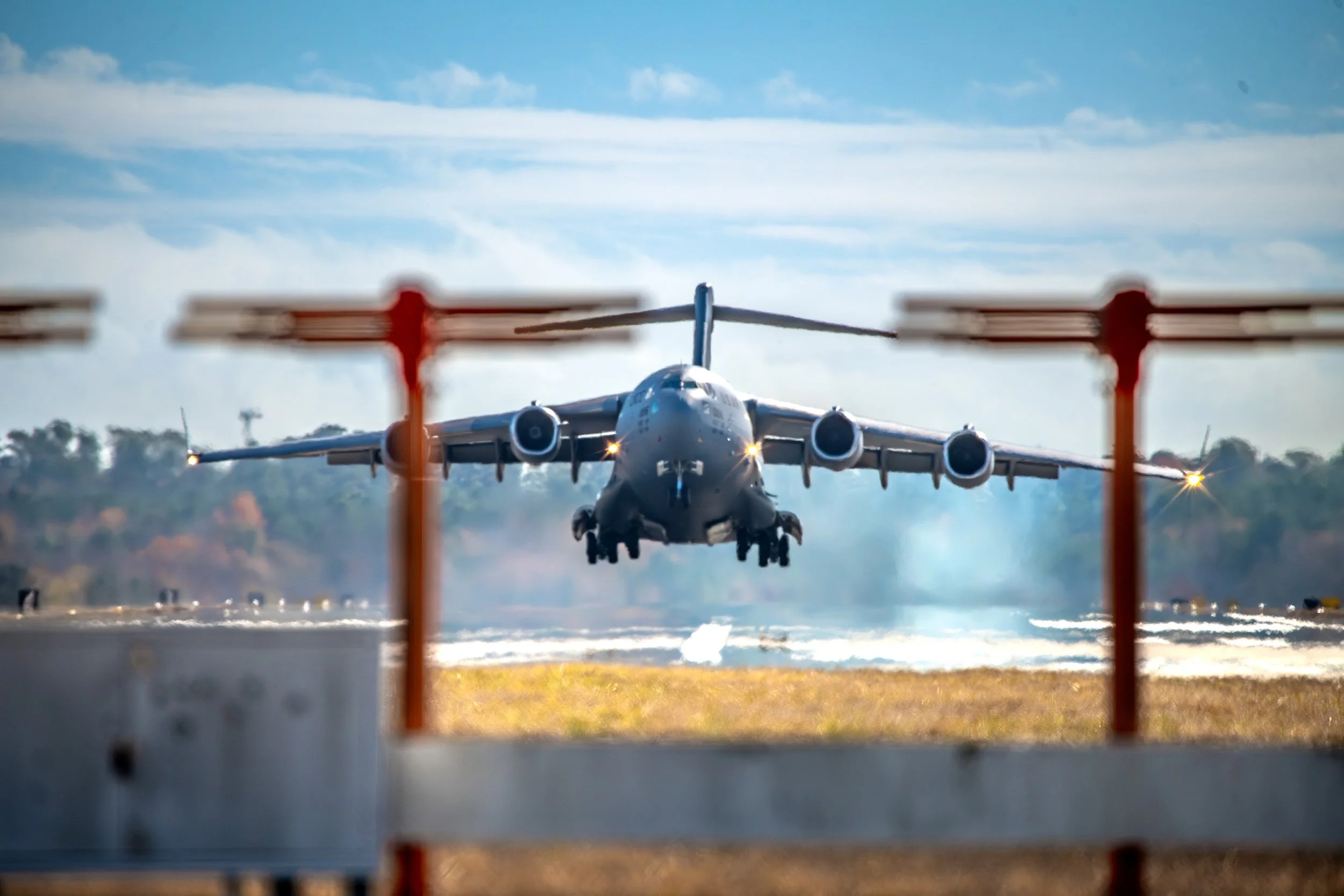 Military cargo airplane taking off from runway, viewed through safety barriers, with cloudy sky and trees in background.