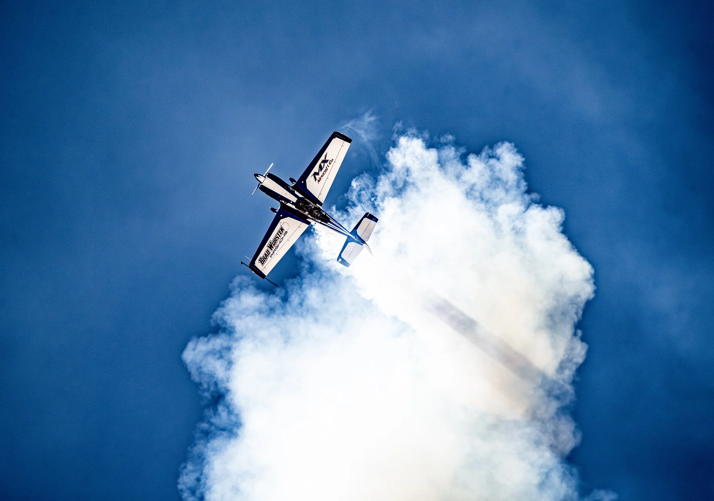 A propeller airplane performing a stunt with a trail of white smoke behind it, flying in a clear blue sky.
