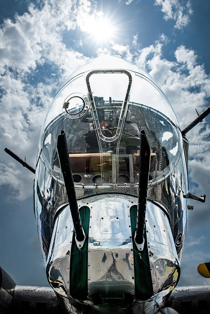 Close-up of the front of a silver military aircraft with a transparent cockpit canopy, looking upward towards a cloudy sky with the sun shining overhead.