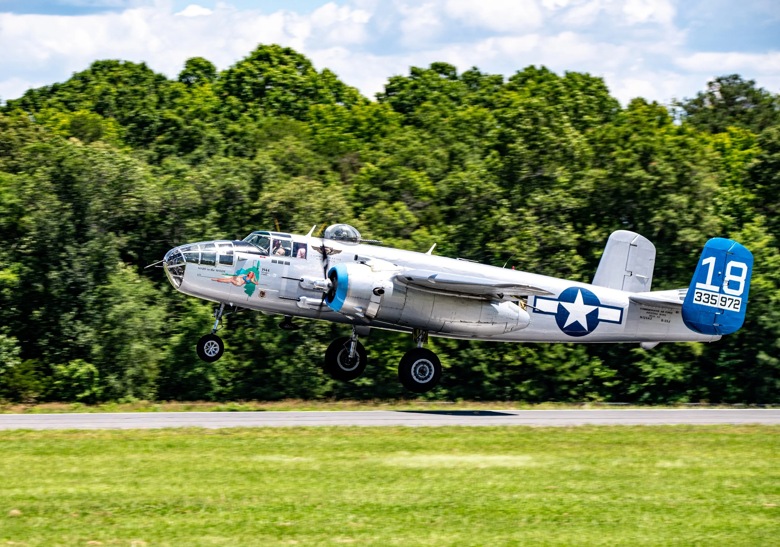 A vintage military aircraft, a North American B-25 Mitchell bomber, taking off from a runway with green trees in the background. The plane has U.S. Army markings and the number 18 on its tail.