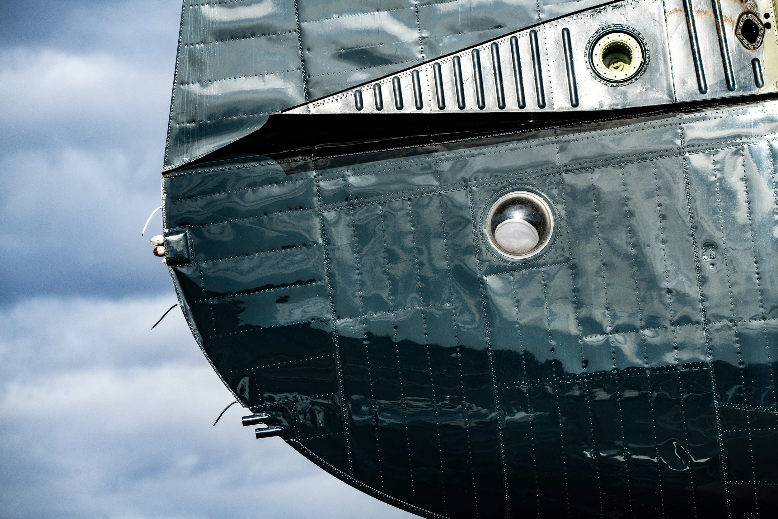 Close-up of the front section of a shiny, metallic aircraft fuselage with rivets, circular windows, and a cloudy sky background.