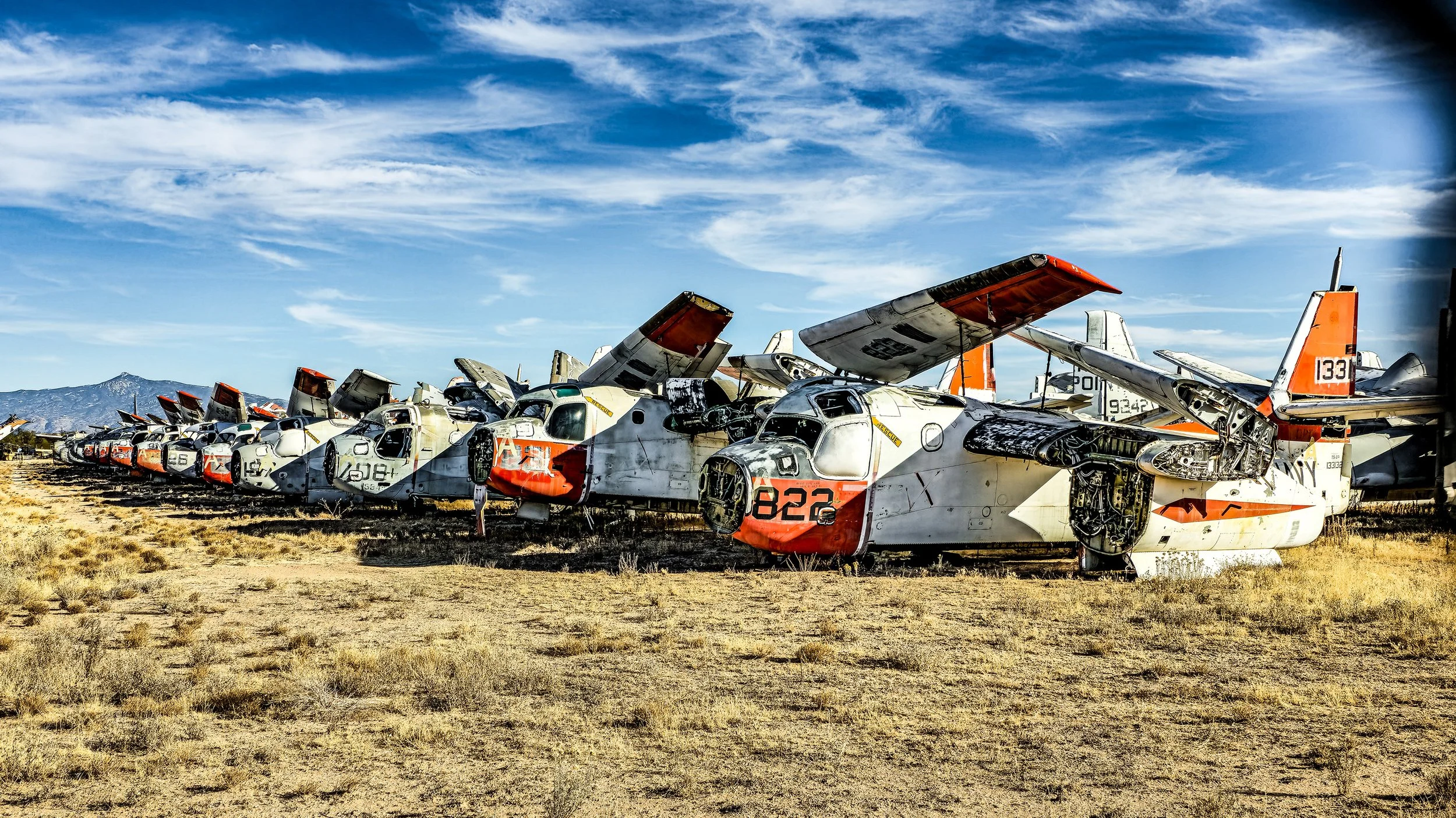 Multiple abandoned and partially disassembled aircraft in a desert landscape under a blue sky with scattered clouds.