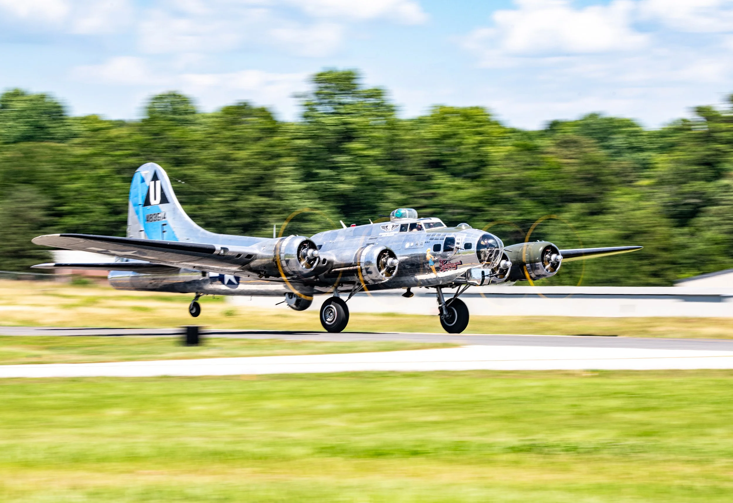 A vintage silver B-17 Flying Fortress airplane taking off from a runway with a blurred green landscape and blue sky in the background.