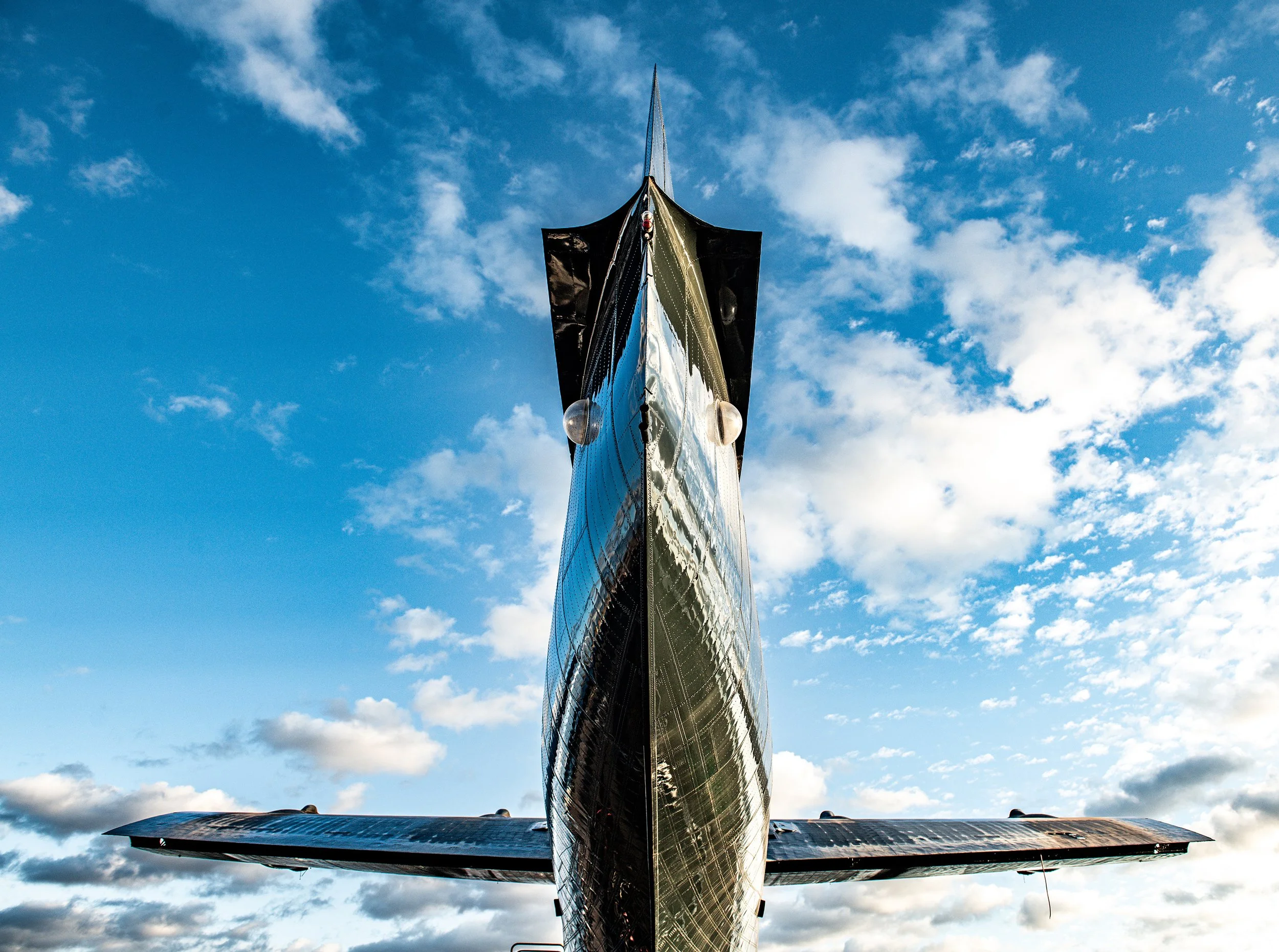 Close-up of the nose of a sleek, black jet aircraft viewed from below, with a partly cloudy sky in the background.