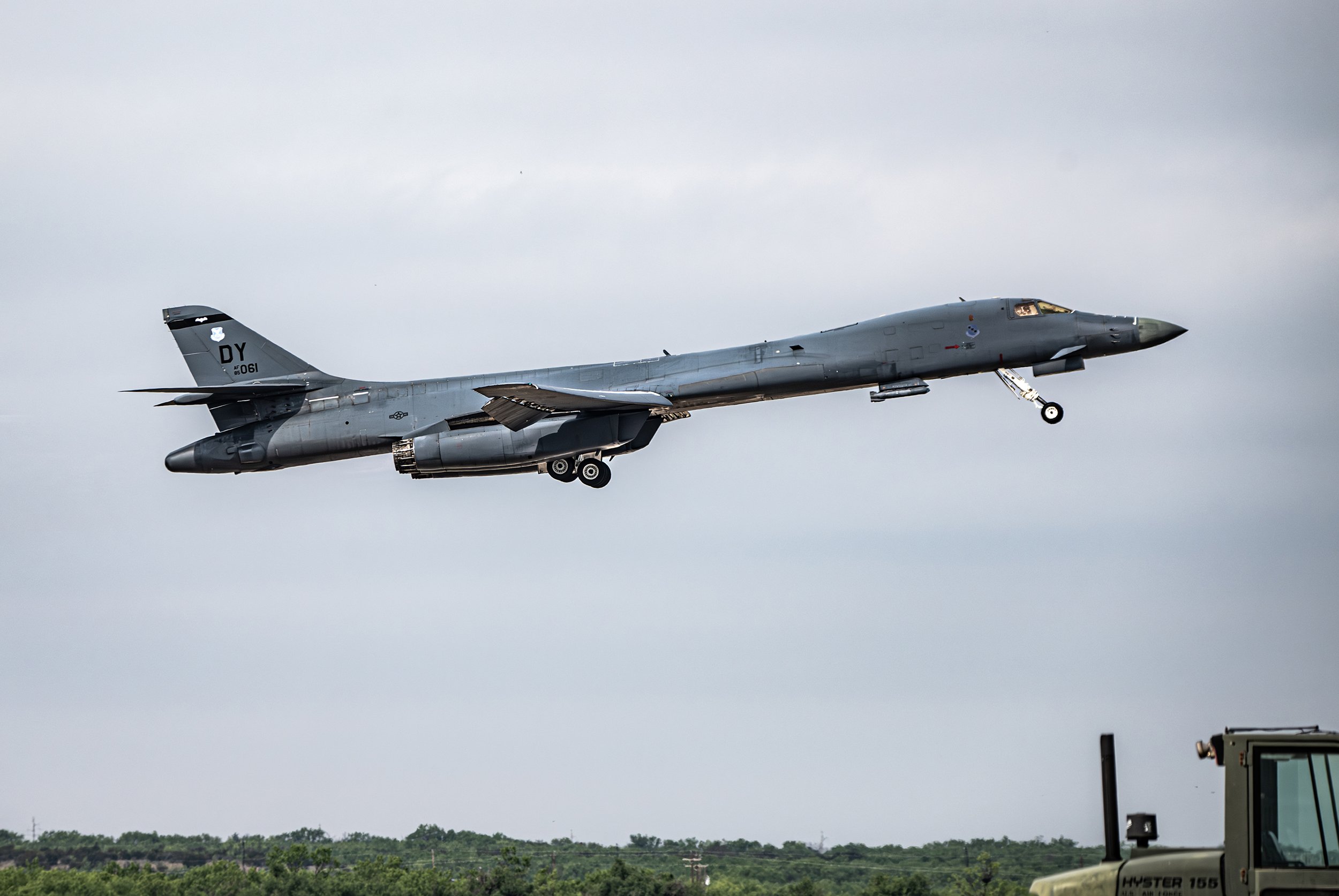 A military fighter jet flying low over a green field, with gray clouds in the sky.