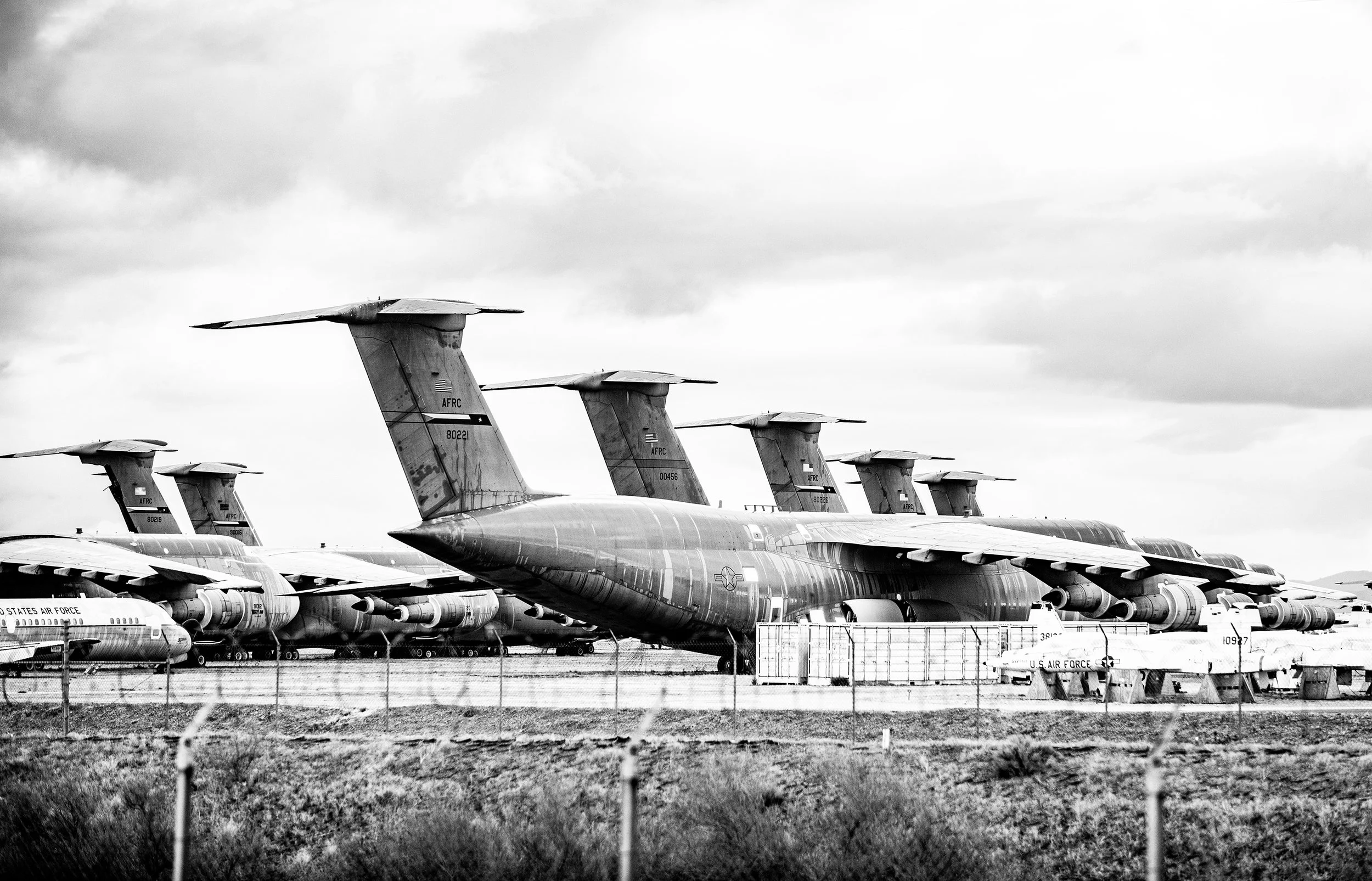 Row of large military cargo planes parked on an airfield, with clouds overhead, in black and white.
