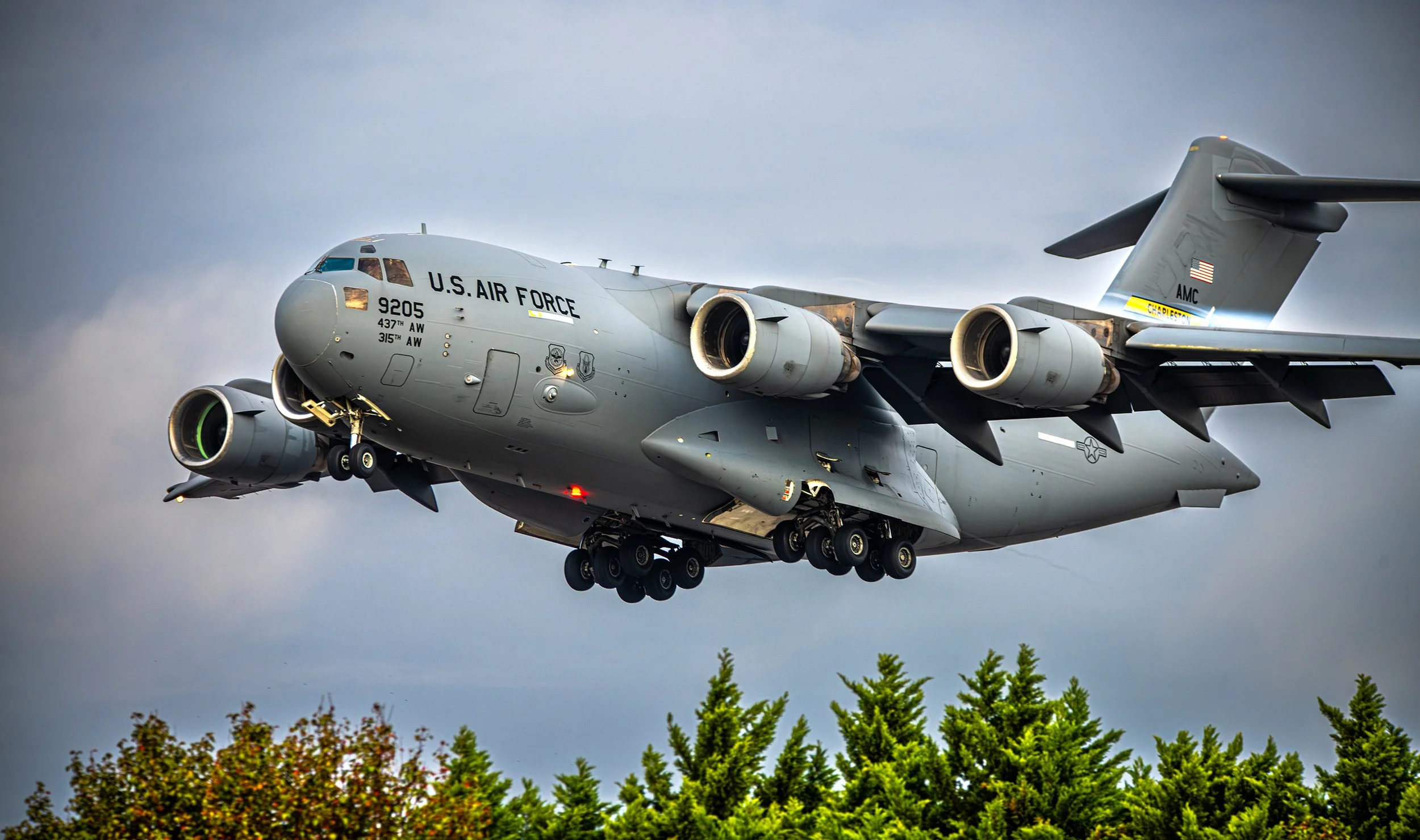 A U.S. Air Force C-17 Globemaster III military cargo aircraft flying low above green trees with gray sky background.