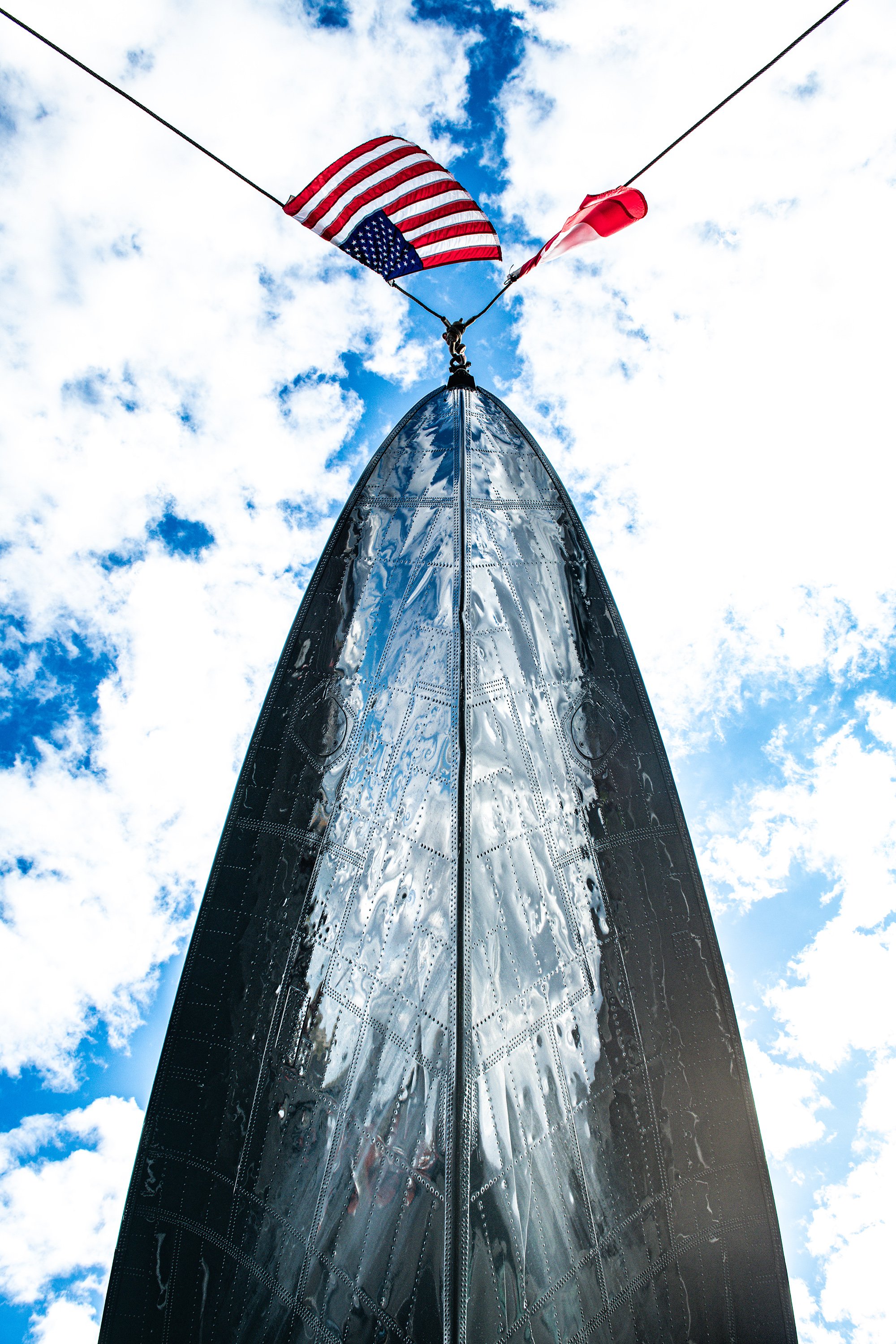 The nose of a shiny, metallic aircraft with an American flag and a red and white flag attached above it, viewed from below against a mostly blue sky with scattered clouds.