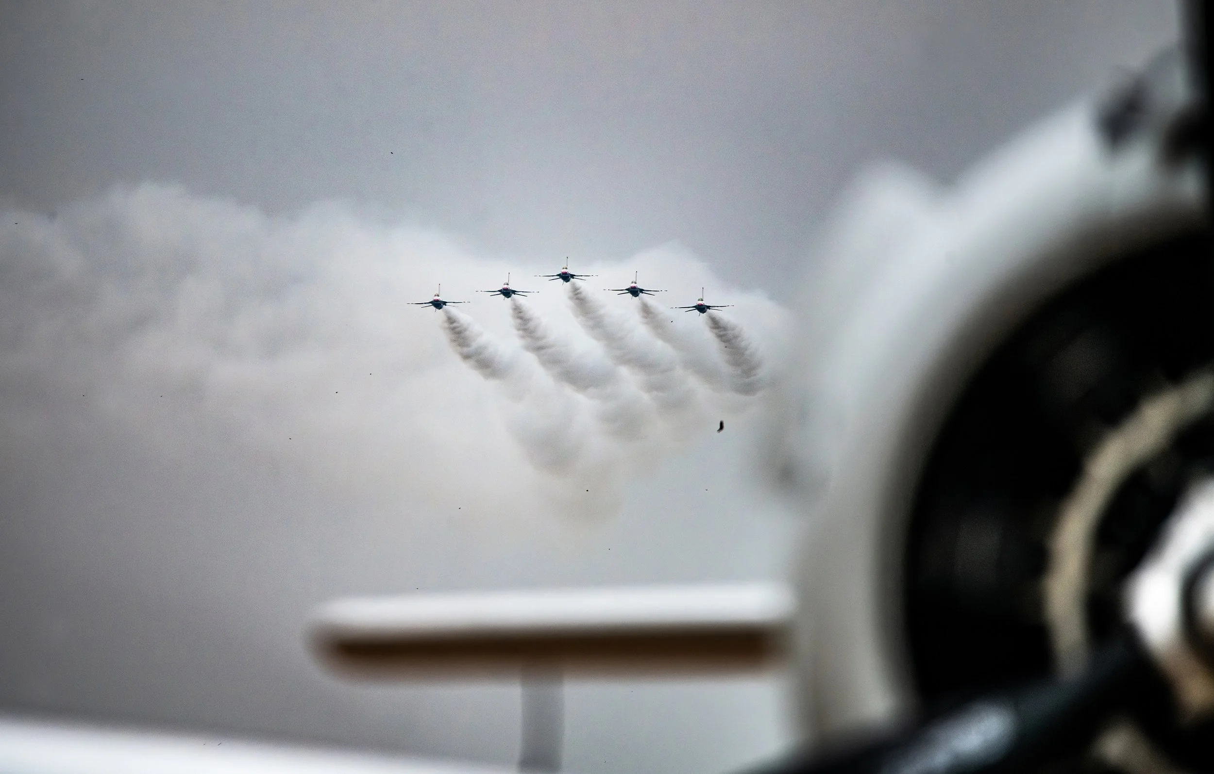 Five airplanes flying in a formation, emitting smoke trails, with the photo taken from inside another aircraft, showing part of the cockpit and an emergency exit sign.