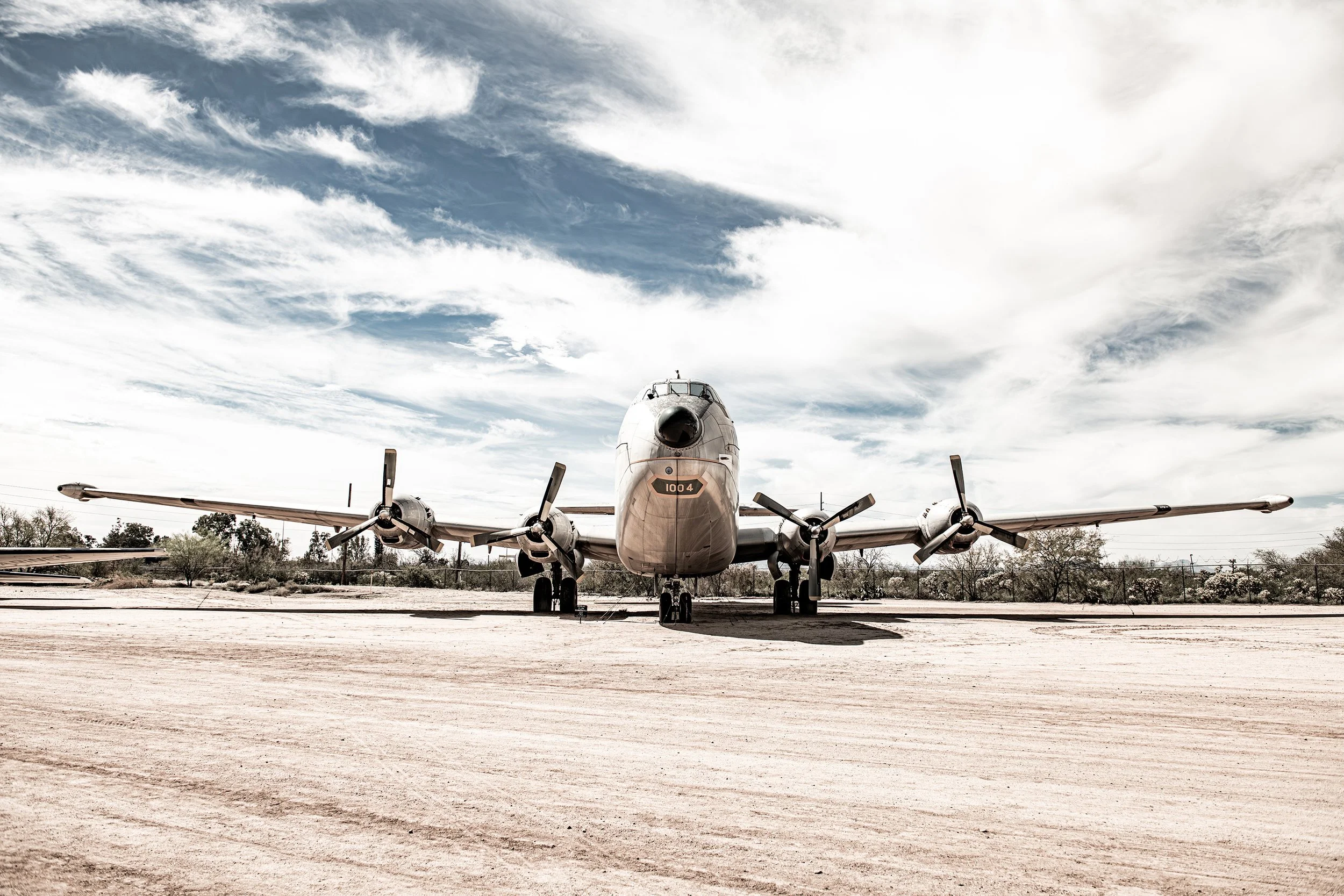 Front view of a large, vintage military propeller plane parked on a dirt airfield under a partly cloudy sky.