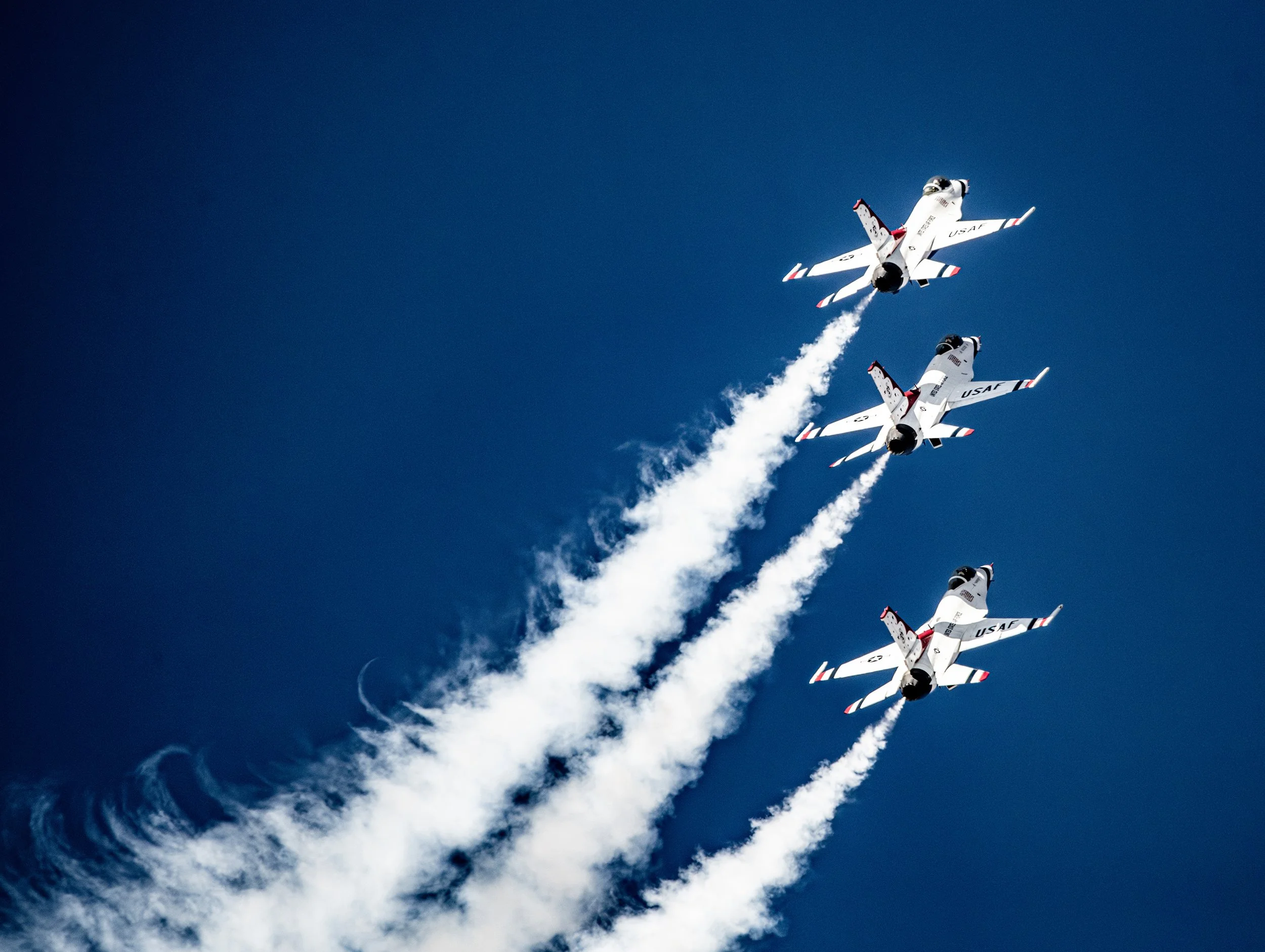 Three fighter jets flying in formation, leaving white smoke trails against a clear blue sky.