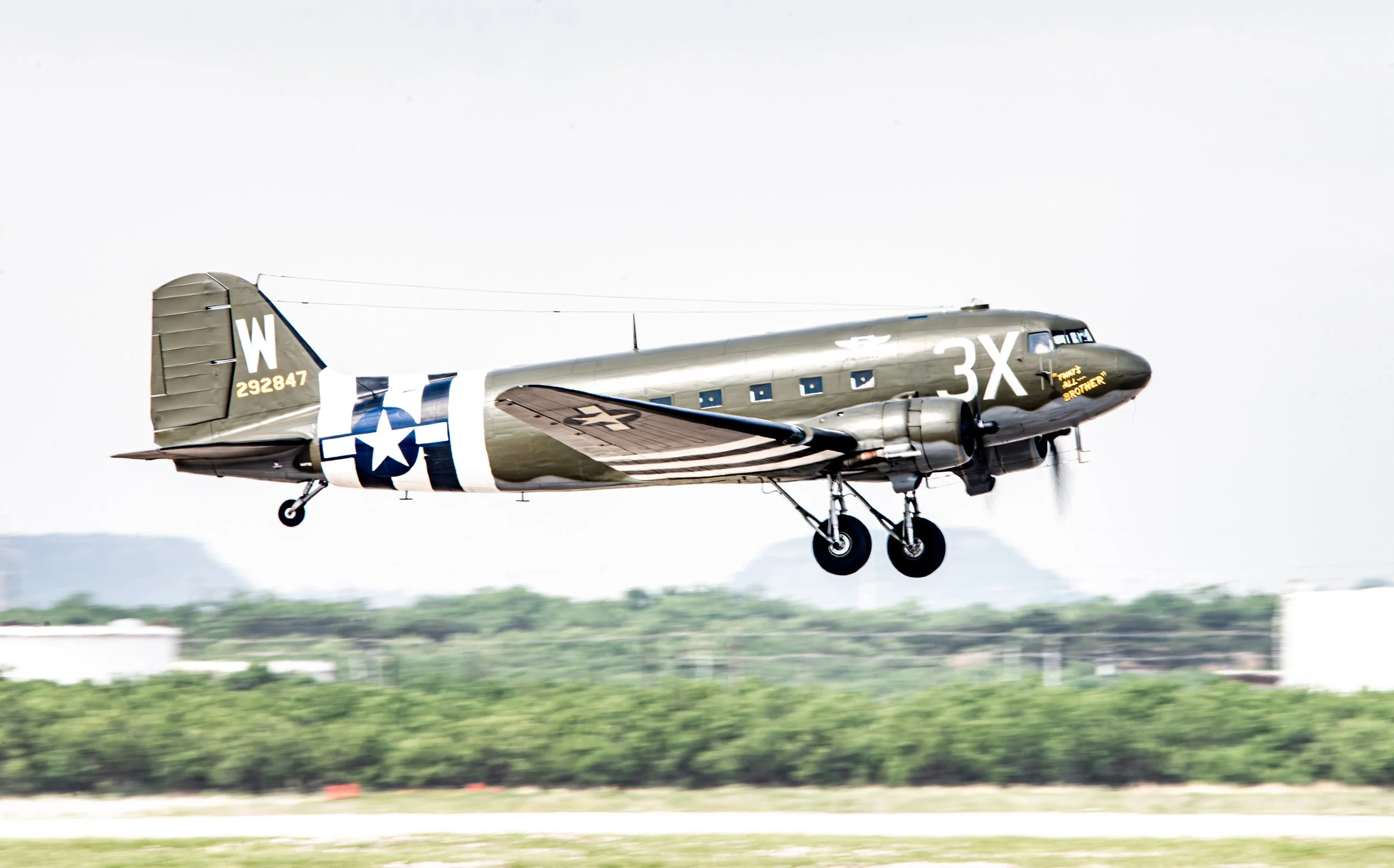 A vintage military aircraft in camouflage colors with U.S. Air Force insignia, landing gear extended, flying low over a field with trees and white buildings in the background.