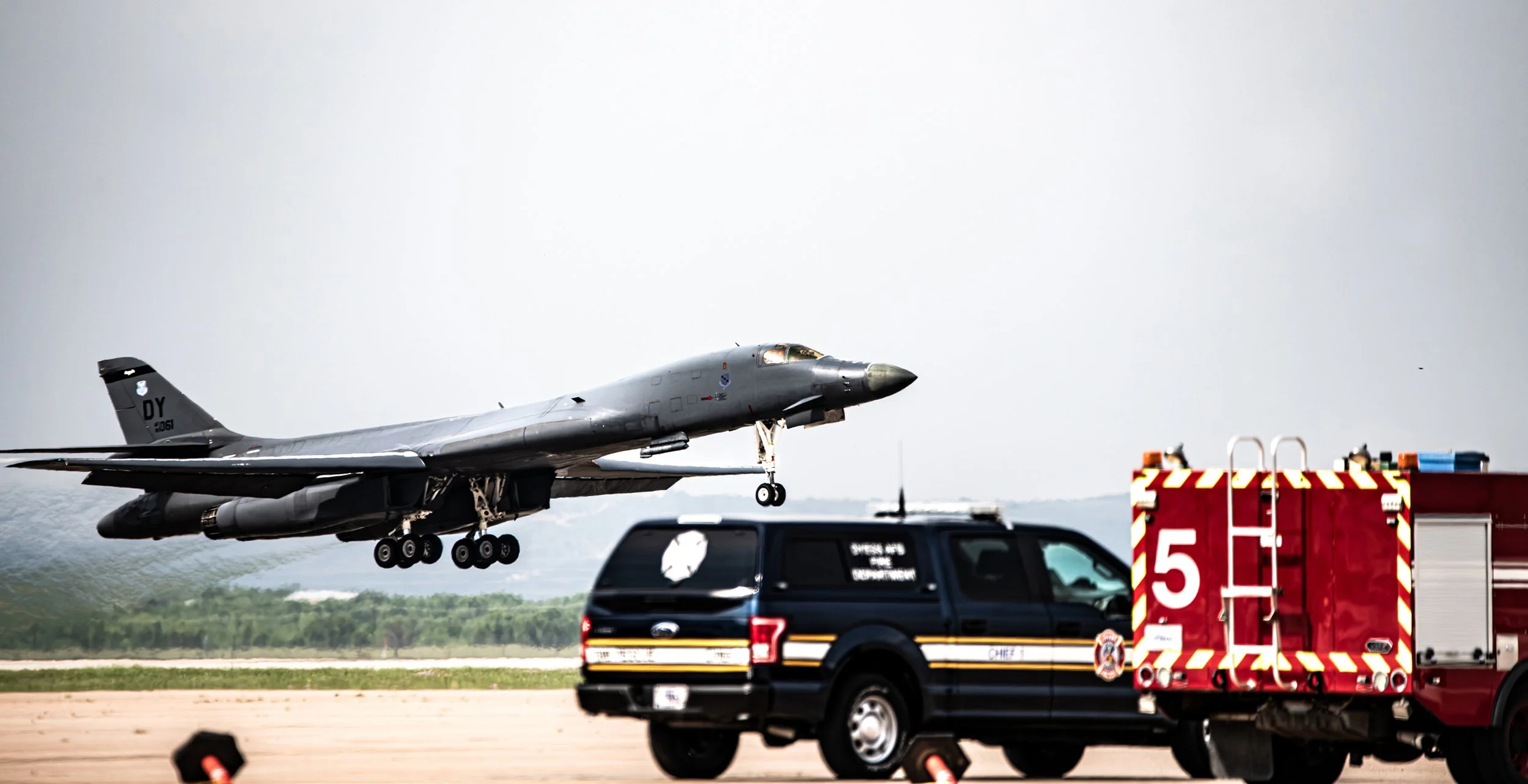 A military jet aircraft taking off near a fire truck and a police vehicle at an airbase.