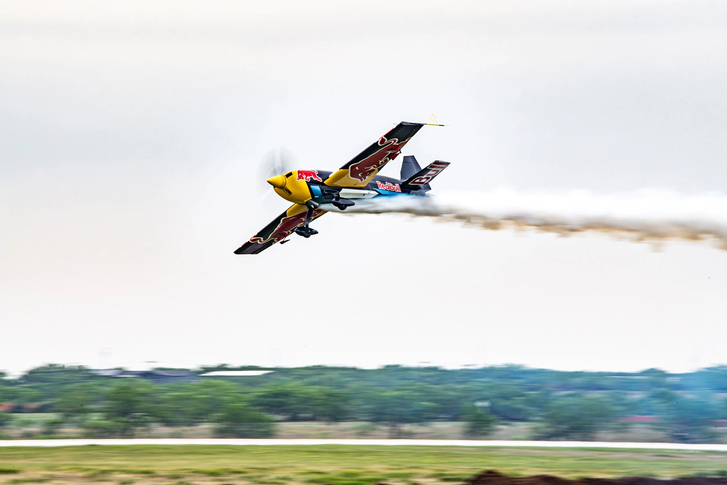A Red Bull racing airplane flying close to the ground, leaving a trail of smoke behind it during an air race or demonstration.