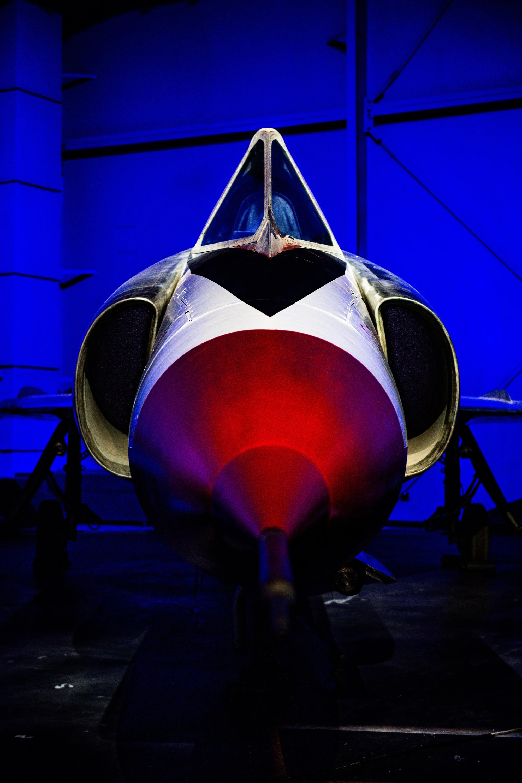 Front view of a fighter jet in a dimly lit hangar with blue lighting, showing the nose and cockpit.