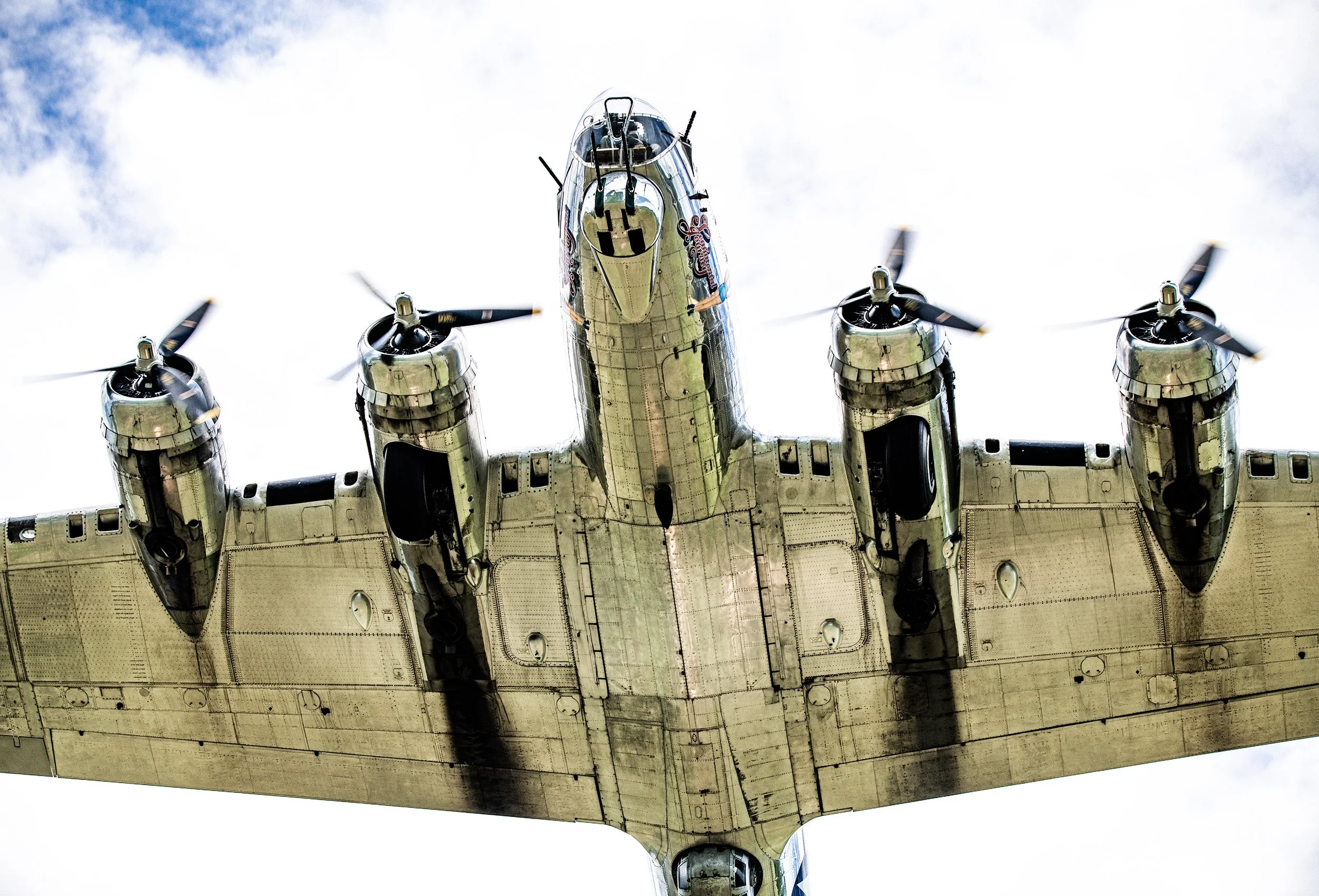 A military aircraft flying overhead, viewed from below, displaying four propellers and a detailed underside.