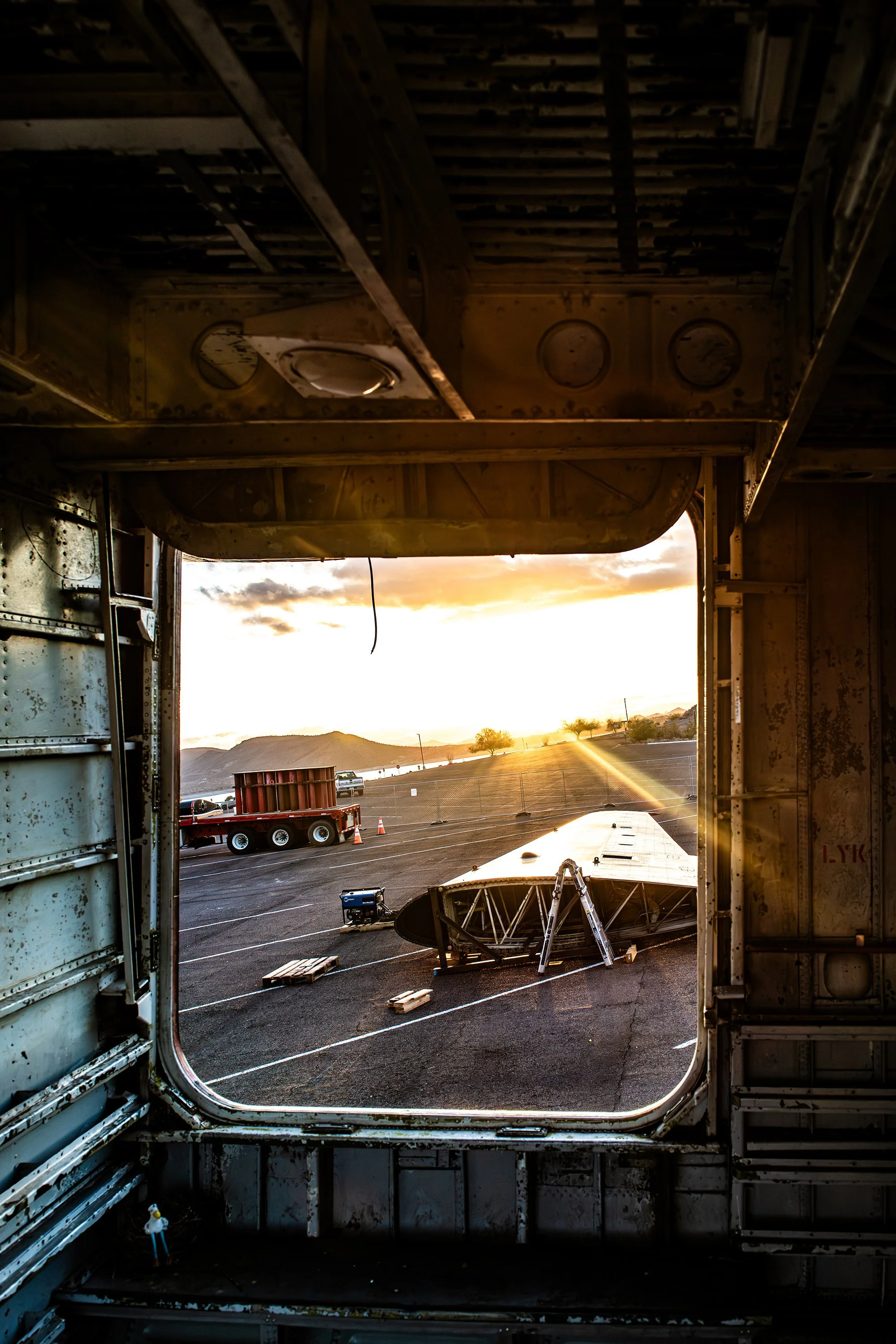 View through the large open cargo door of an aircraft showing the wreckage of the airplane's wing on the tarmac with a trailer, small tools, and equipment around it, and a sunset in the background.