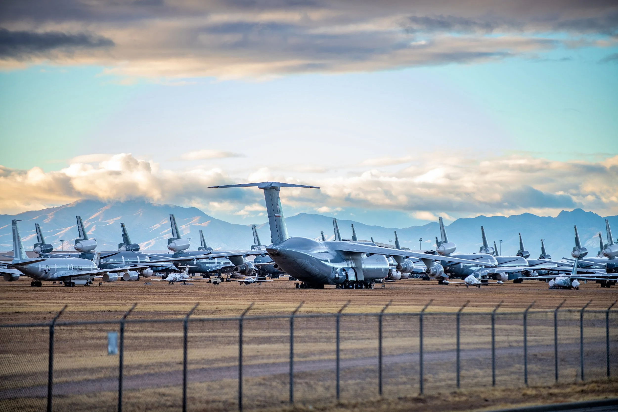 Military cargo planes and fighter jets parked on a tarmac, with mountains and a cloudy sky in the background.