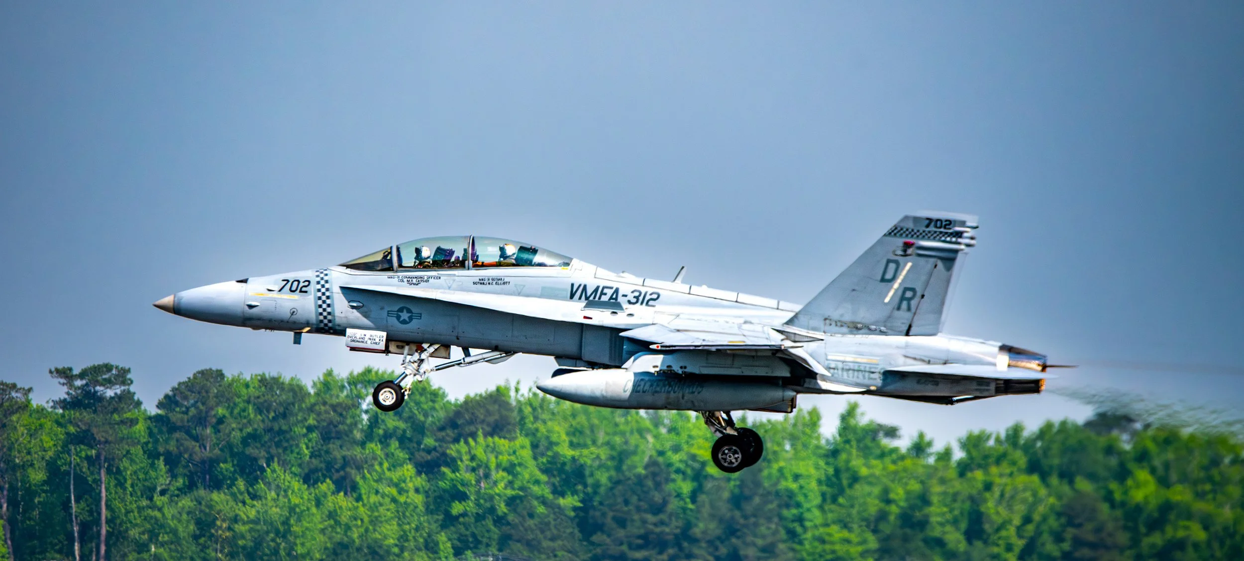 A jet fighter aircraft flying low over green trees, with landing gear extended.