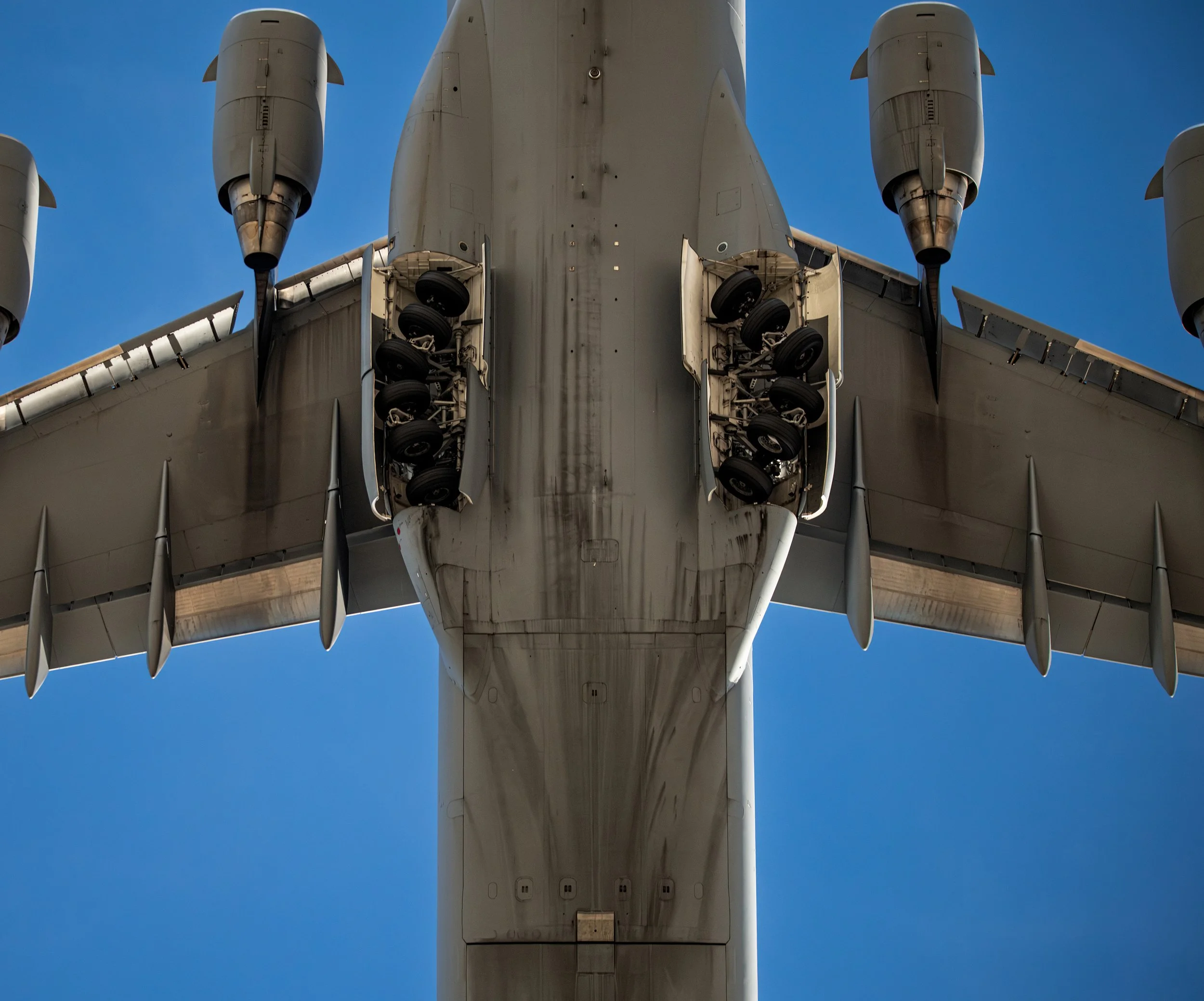 Close-up view of the underside of a large military cargo airplane showing landing gear compartments with wheels, against a clear blue sky.