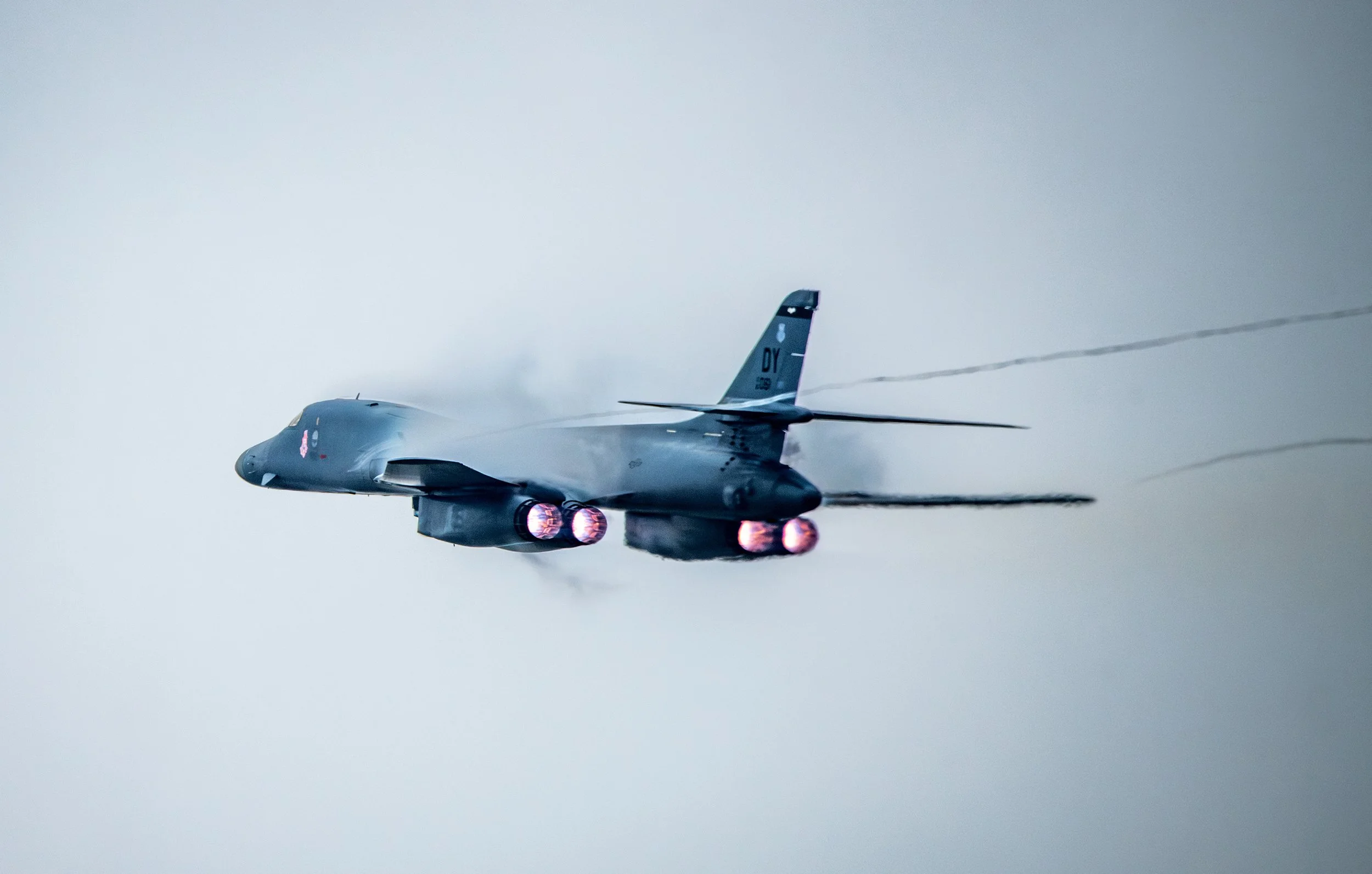 Fighter jet airplane flying through the sky with afterburners on, emitting bright flames and leaving black smoke trails.
