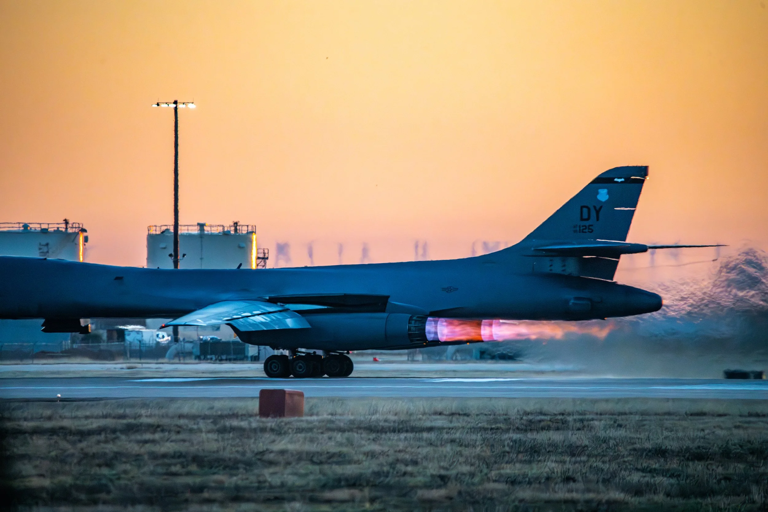 Military jet aircraft on runway with afterburner flame, against a setting sun sky, with fuel tanks and industrial structures in background.