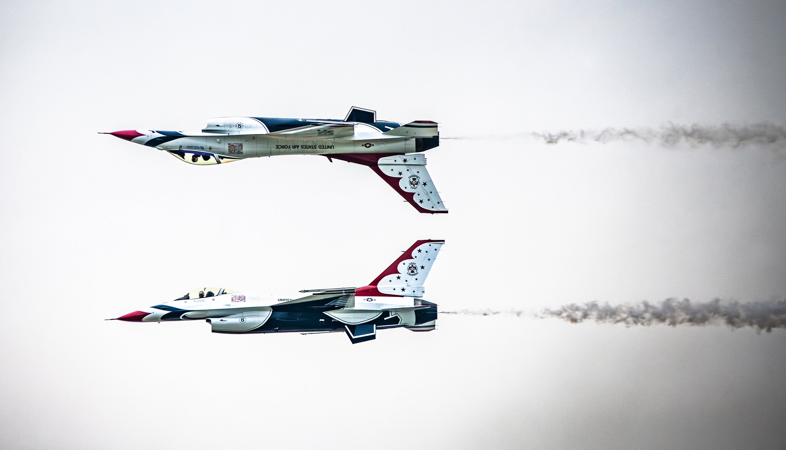 Two United States Navy Blue Angels fighter jets flying upside down in close formation, with smoke trails, against a cloudy sky.