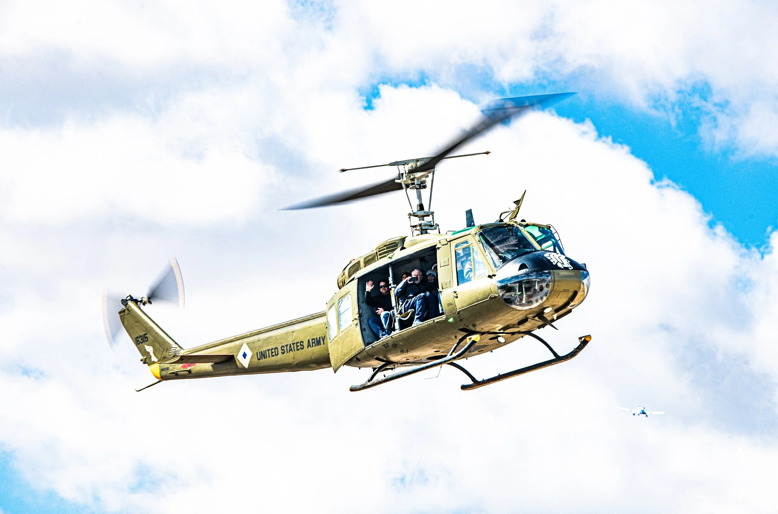 A United States Army helicopter flying in the sky with two people inside, one waving, with clouds and another aircraft in the background.