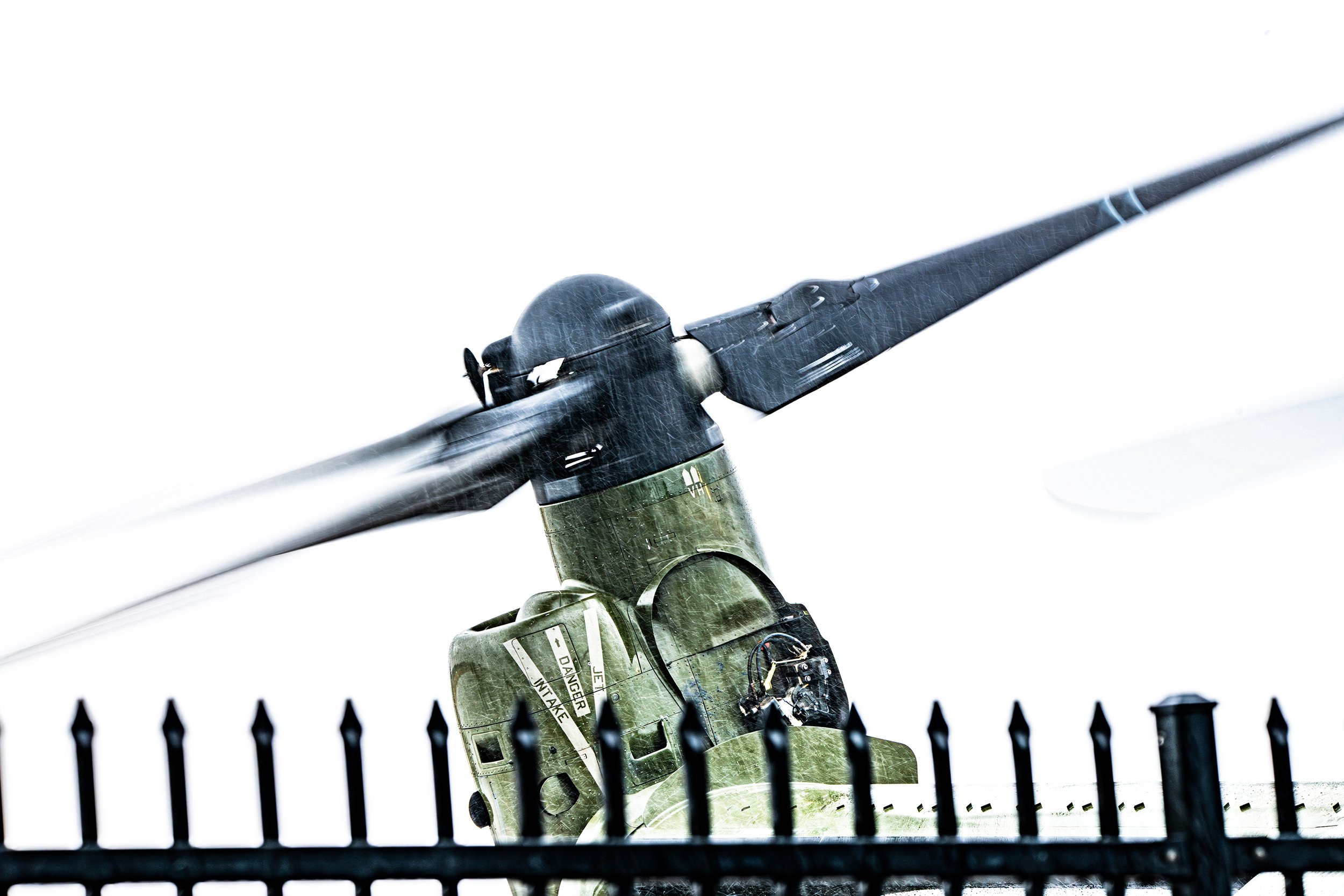 Close-up of a military helicopter's rotor blades in motion, rain falling, with a black fence in the foreground.