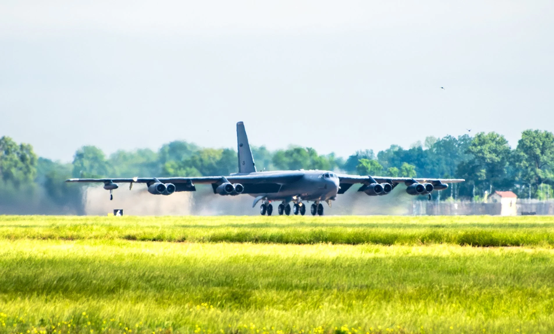 A large military cargo plane landing on a grassy airfield with trees and a small building in the background.