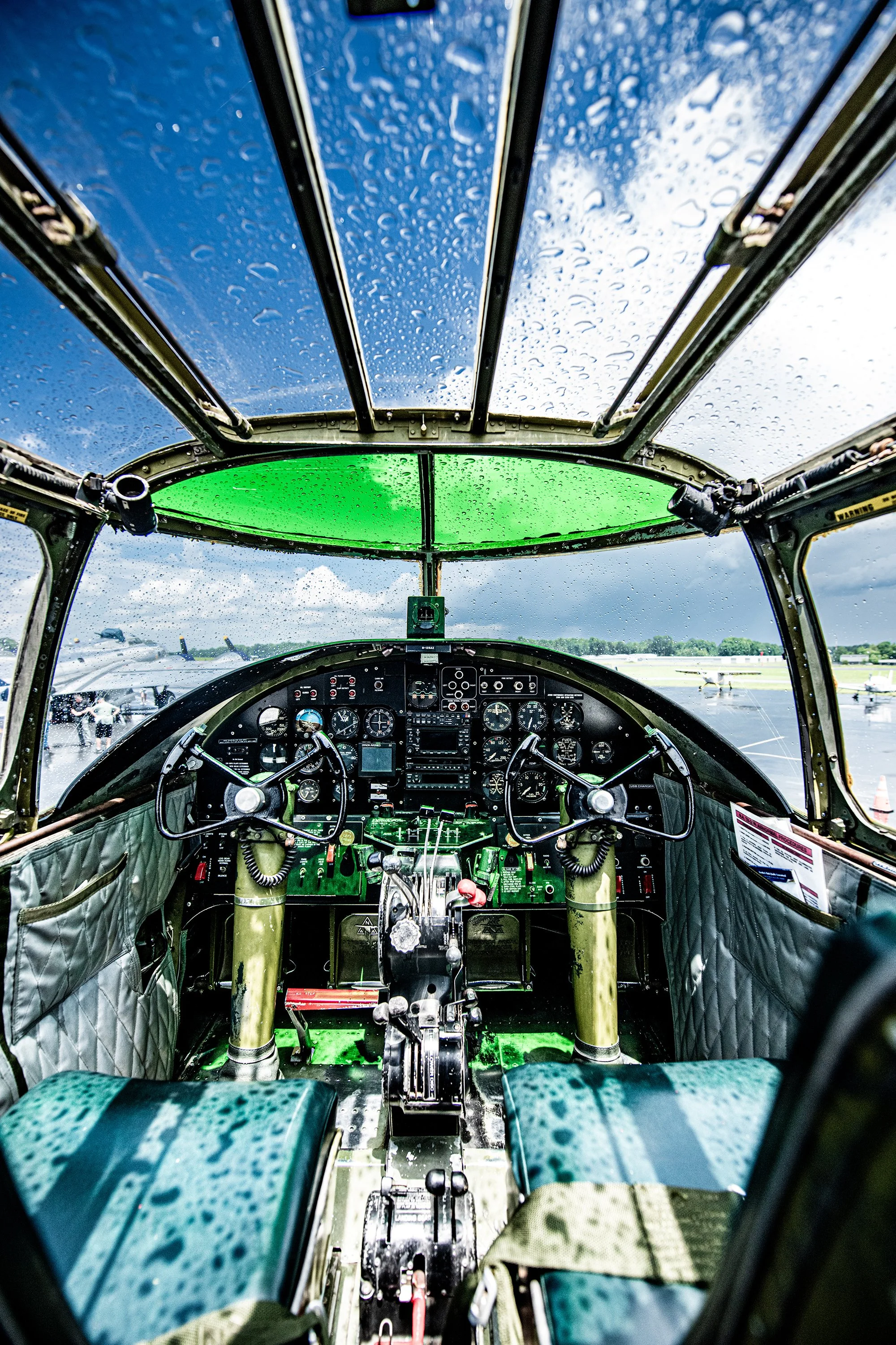 Inside the cockpit of a vintage airplane, rain droplets on the transparent canopy, with an airfield and other planes visible outside.