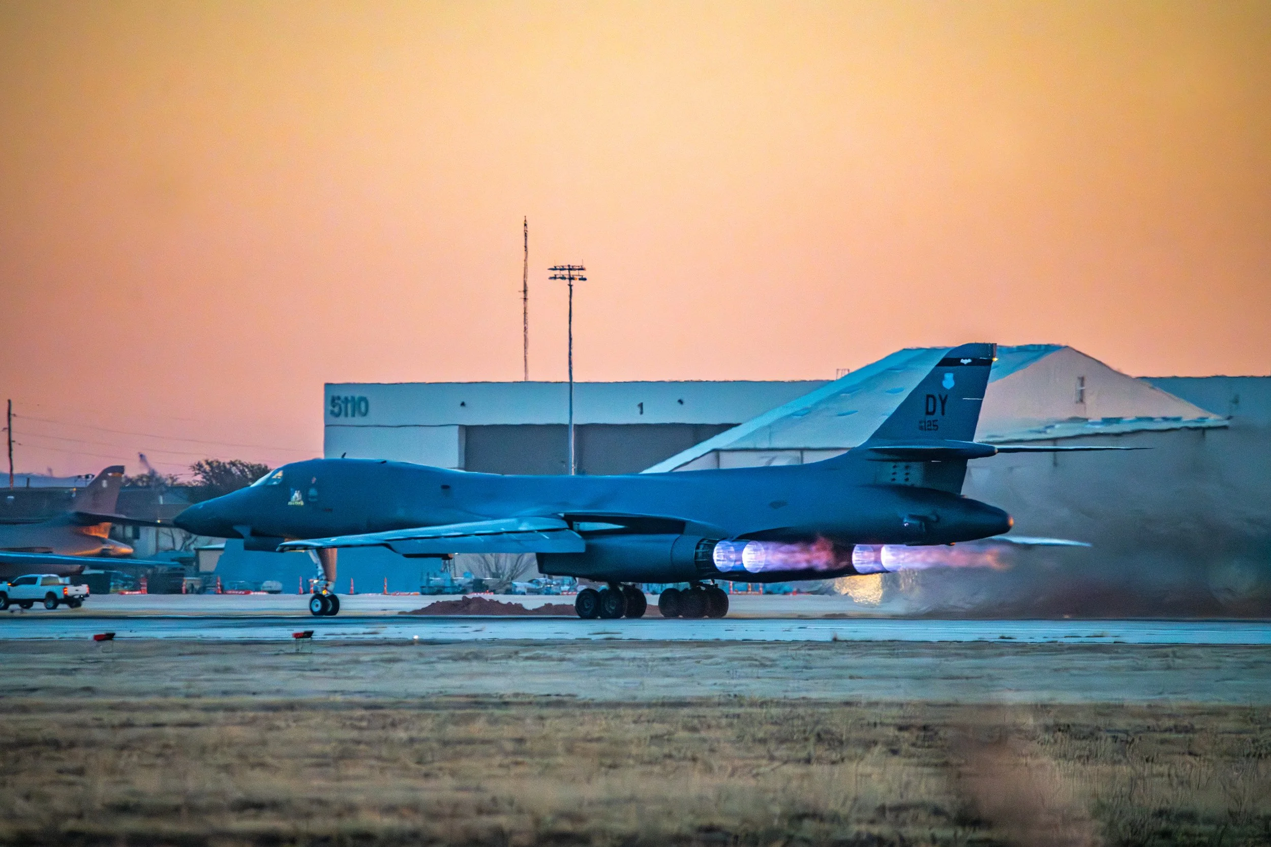 A military jet aircraft on the runway during sunset, with flames visible from its engines.