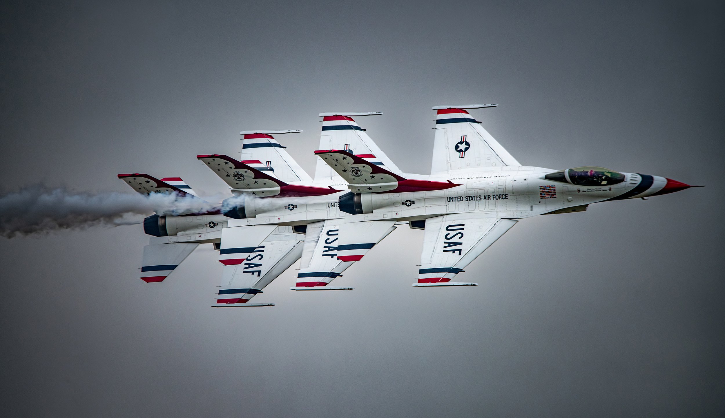 Four United States Air Force fighter jets flying in close formation through a cloudy sky, with visible exhaust smoke trailing behind one jet.