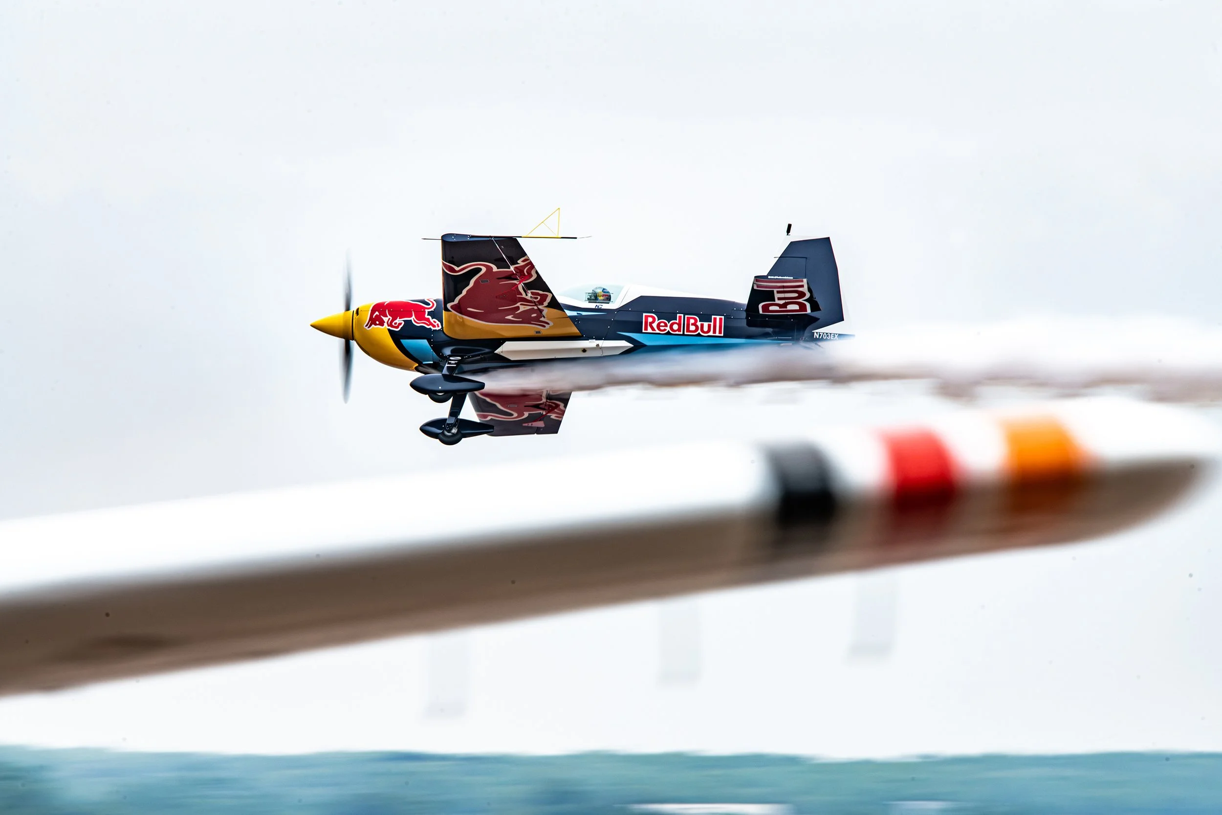 A model jet plane with Red Bull and Red Bull logo design flying close to the camera over the water with a blurred background.