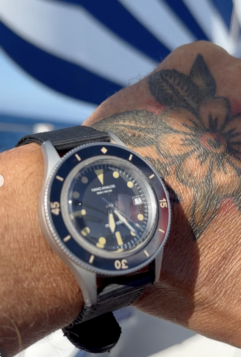 A wristwatch with a black dial and yellow markers, worn on a tattooed wrist, with a background of a blue and white striped umbrella or parasol.