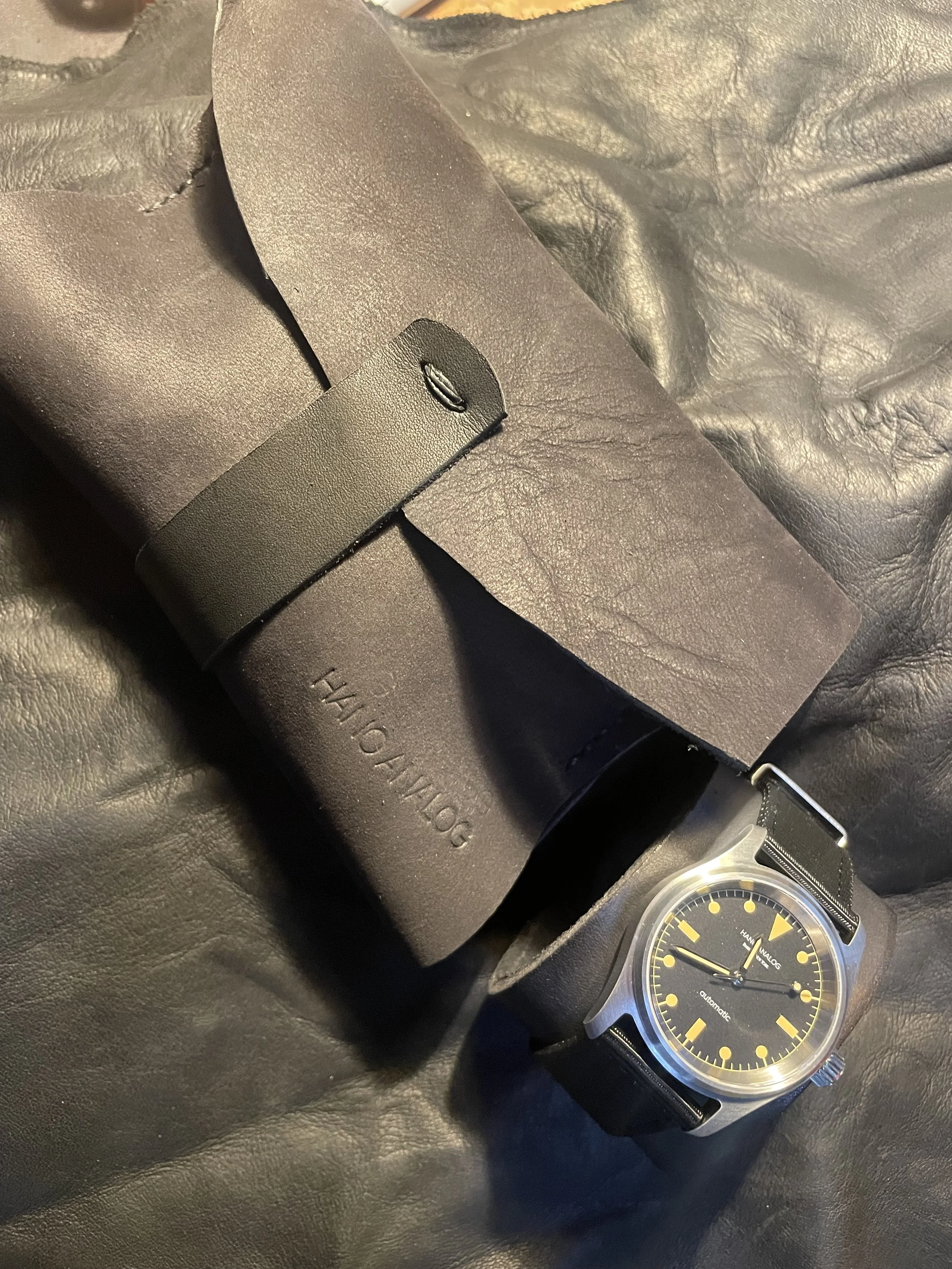 A black wristwatch with a yellow and black dial, a leather wallet, and a black leather bag on a dark surface.