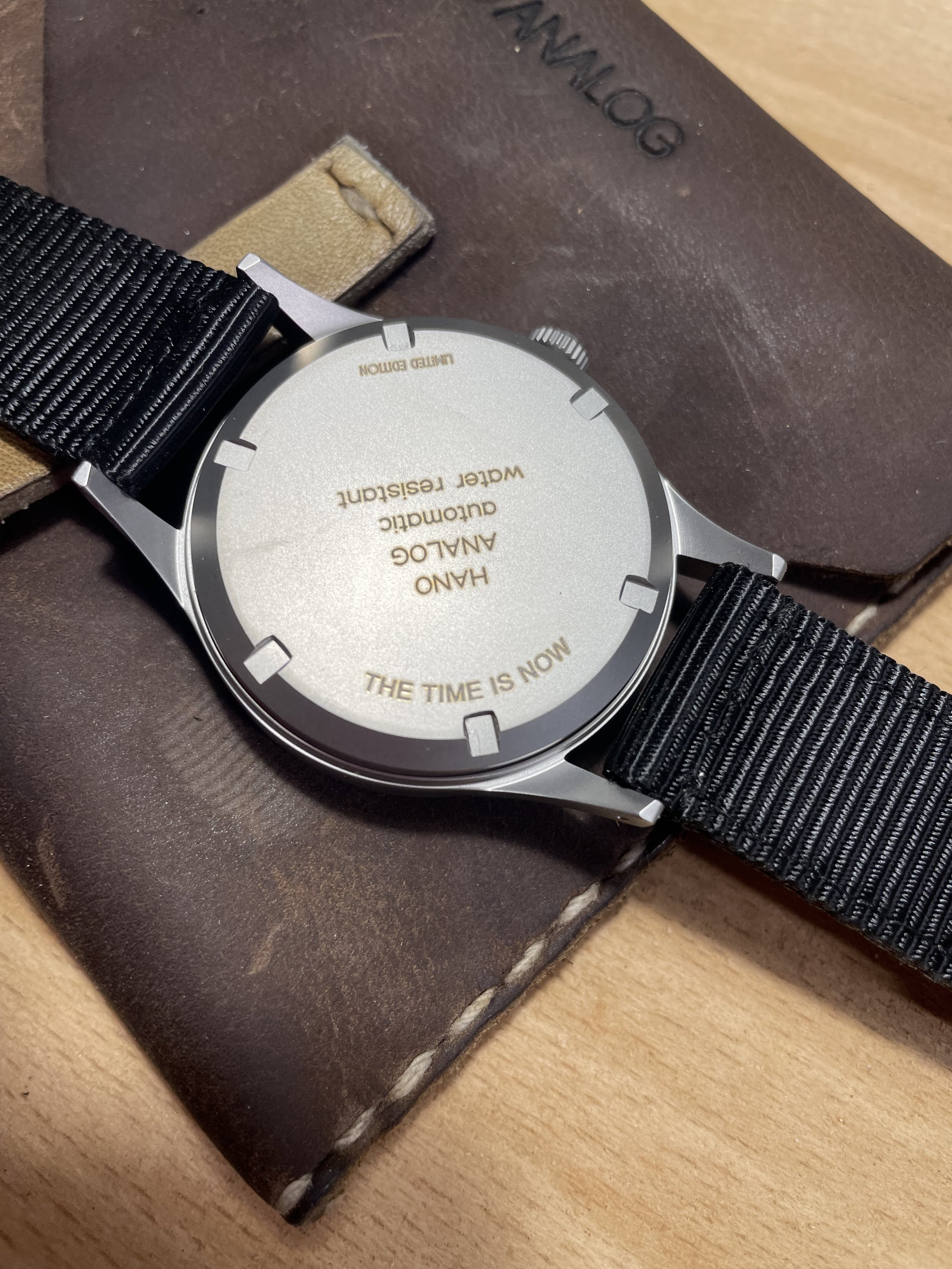 Back of a wristwatch with a silver case, black strap, and a message engraved on the case back that says 'HAND AUDOMATIC WATER RESISTANT THE TIME IS NOW' while the watch is placed on a brown leather item with the word 'NOMLOG' embossed on it.