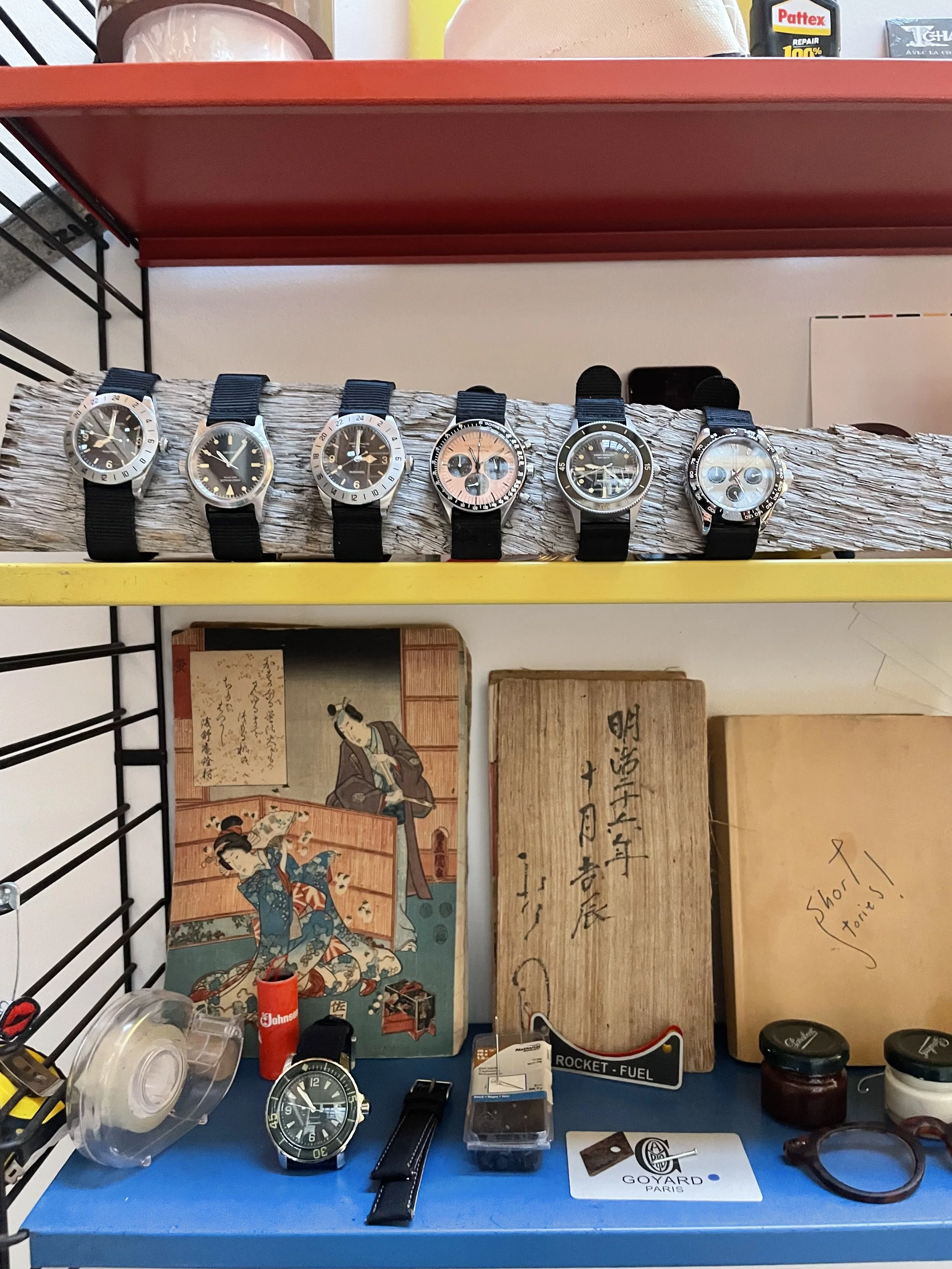 Display of six wristwatches on a wooden log and a blue surface with various small items, posters, and wooden plaques in the background.
