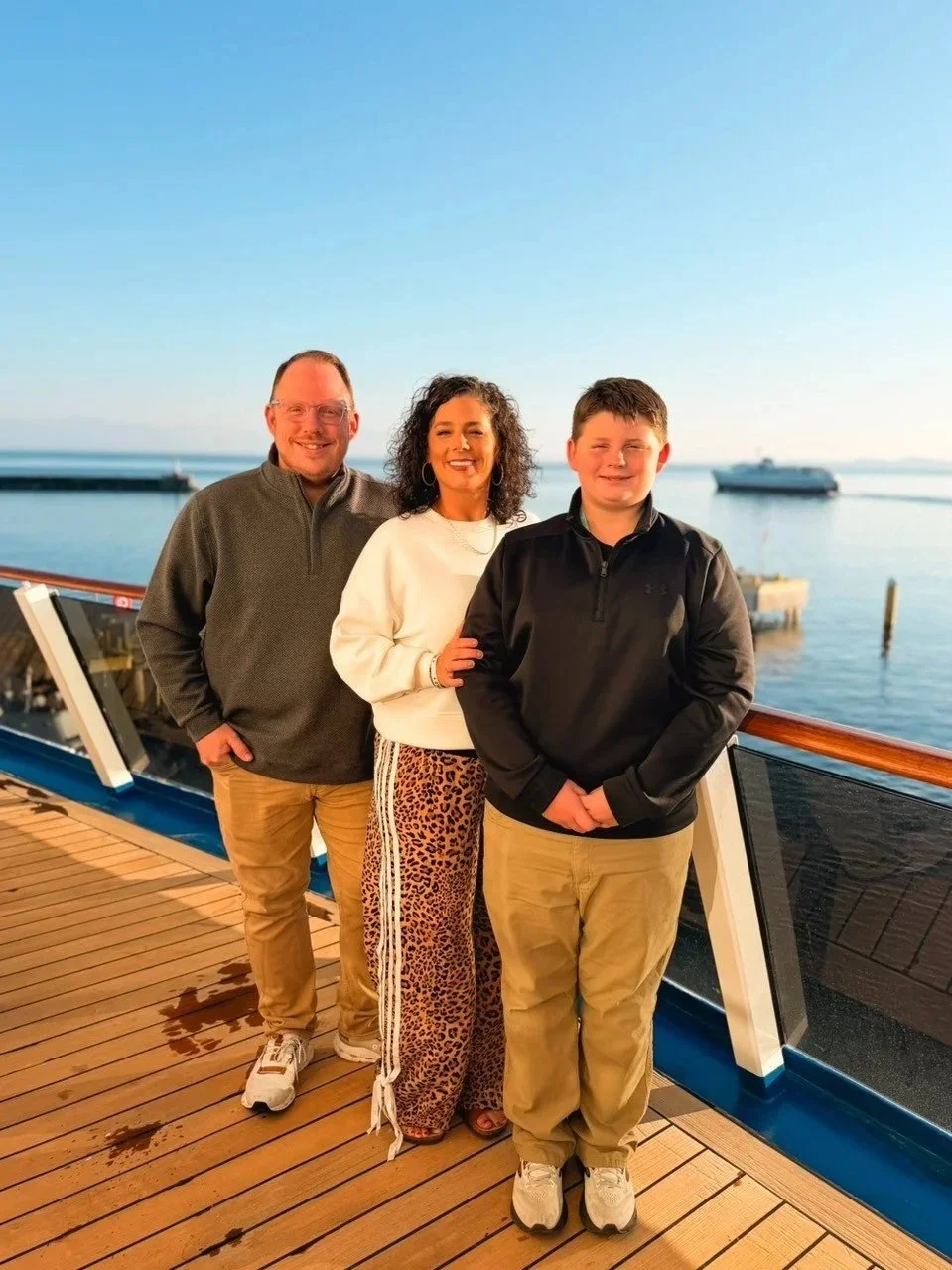 A family of three posing on a ship's deck with a view of the water and boats in the background during sunset.
