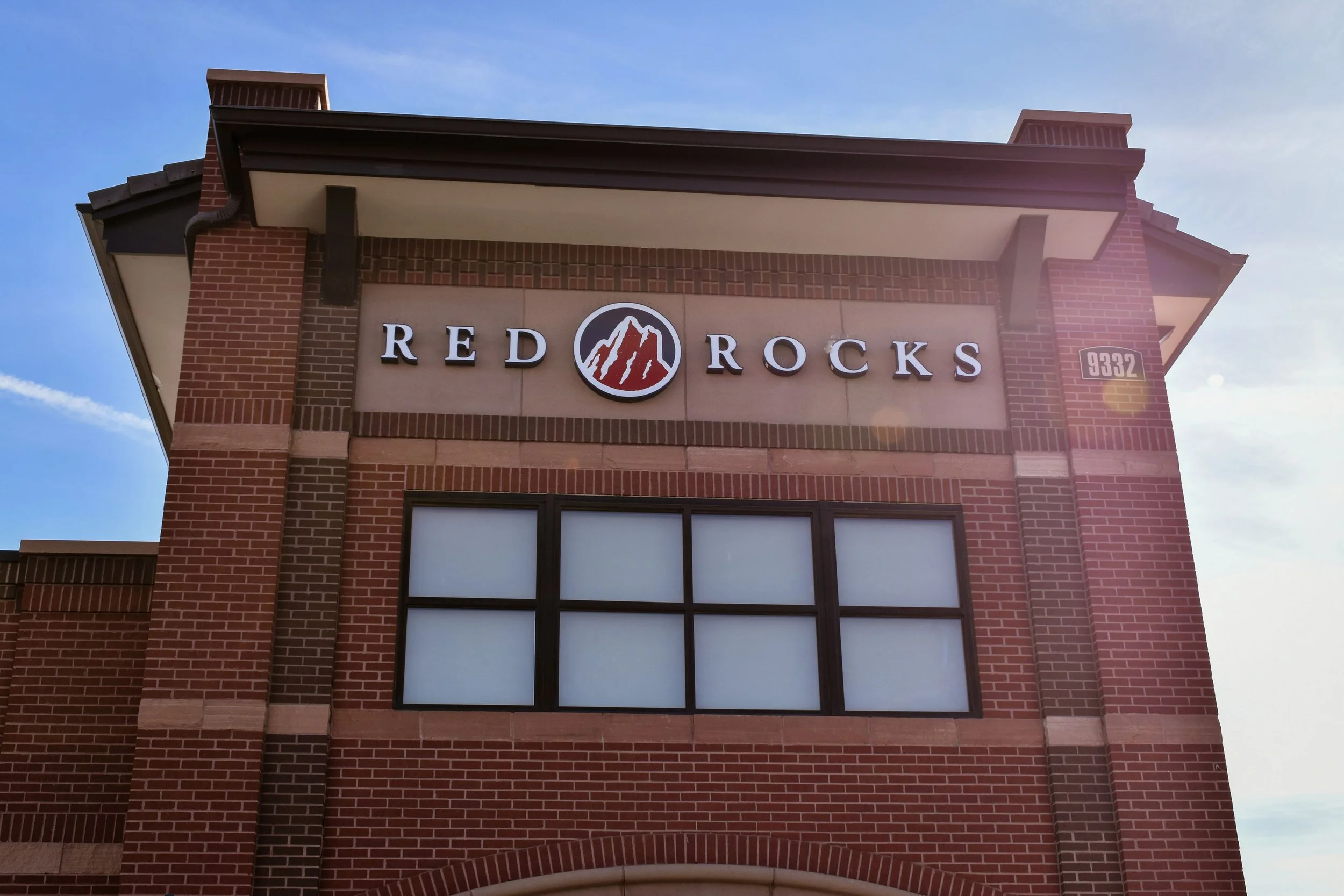 The exterior of a brick building with a sign that reads 'Red Rocks' and includes a logo of a mountain with red rock formations. The building's address, 9332, is visible on a plaque on the right side.