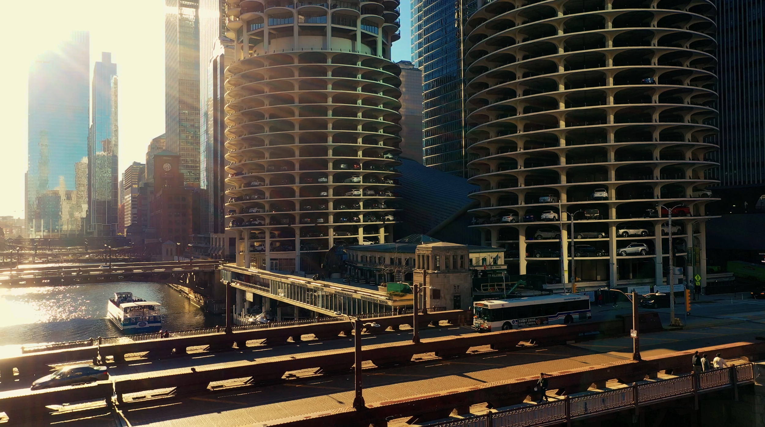 Cityscape with tall buildings, a river with boats, a bridge, and a parking garage with rounded architecture in the background during sunset.
