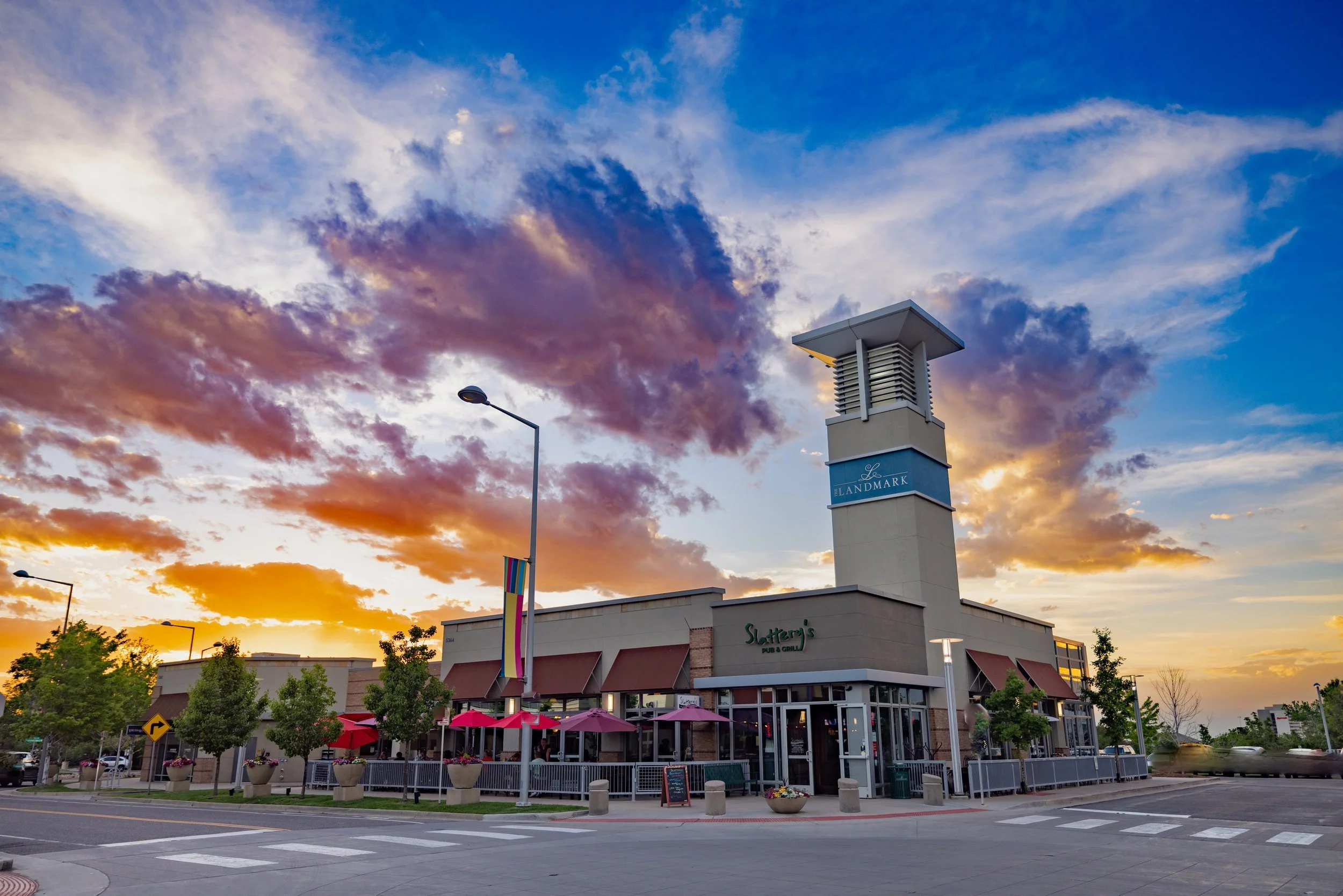 A shopping plaza during sunset with a colorful sky, featuring a light-colored building with outdoor seating, red umbrellas, and a tall tower with signs for Landmark and Slattery's Pub & Grill.