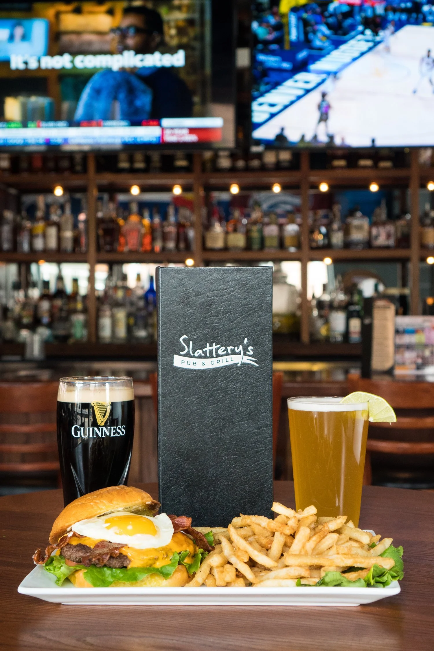 A meal at a pub features a burger with bacon, lettuce, sunny-side-up egg, French fries, a pint of Guinness beer, and a glass of pale beer garnished with lemon, with a black menu standing upright on the table and a bar with liquor bottles in the background.