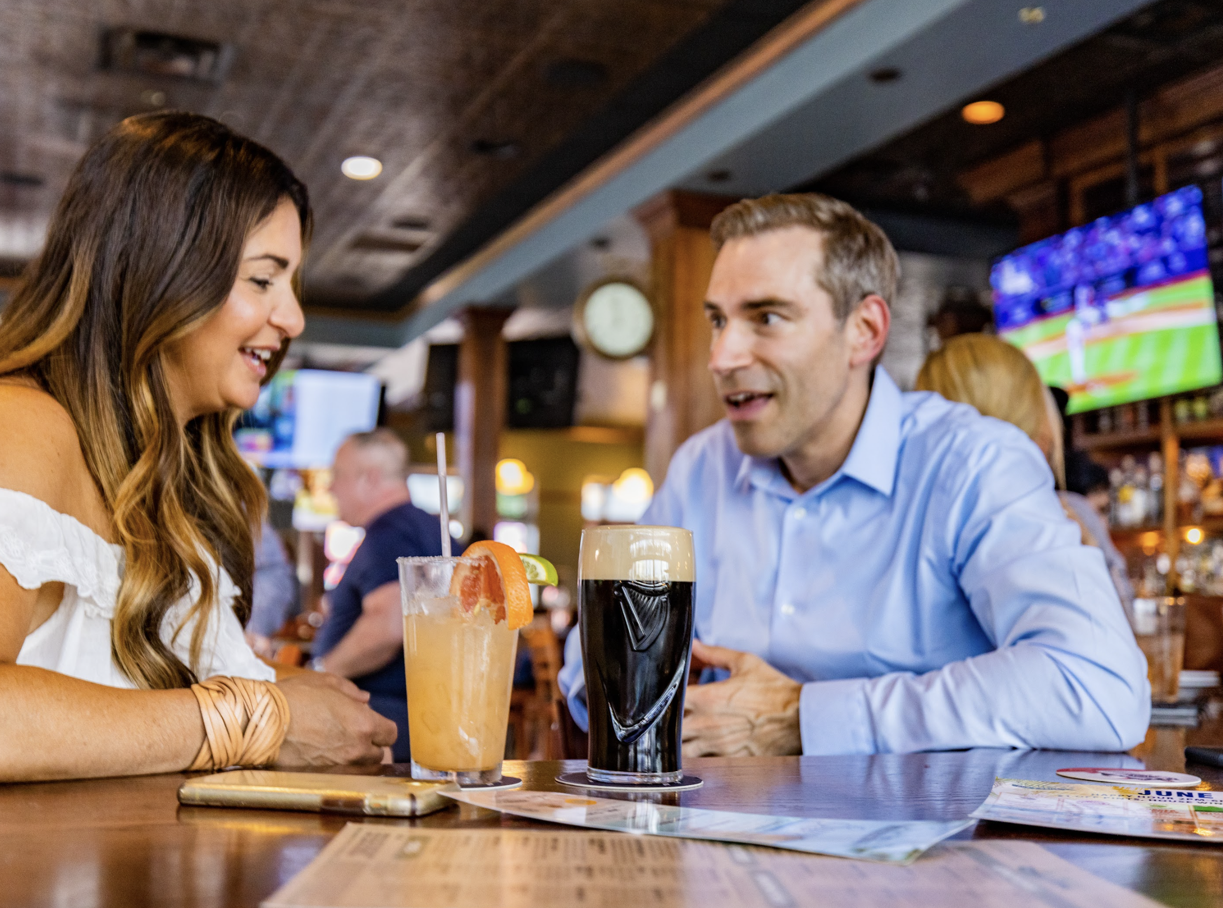 A man and a woman sitting at a bar table, engaged in conversation. The woman has long brown hair and is smiling, while the man has short hair and is wearing a light blue shirt. They have drinks in front of them, one appears to be a cocktail with citrus garnishes, and the other is a dark stout beer. The background shows a lively bar environment with a flat-screen TV, other patrons, and warm lighting.