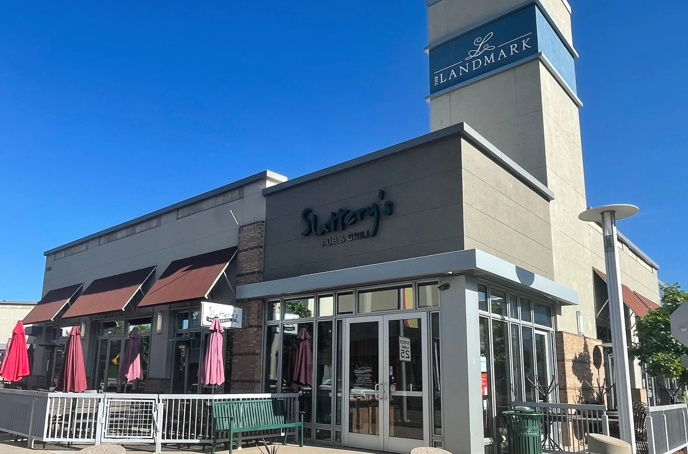 Exterior of Slattery's Pub & Grill with outdoor seating and umbrellas, located in The Landmark shopping center, under a clear blue sky.
